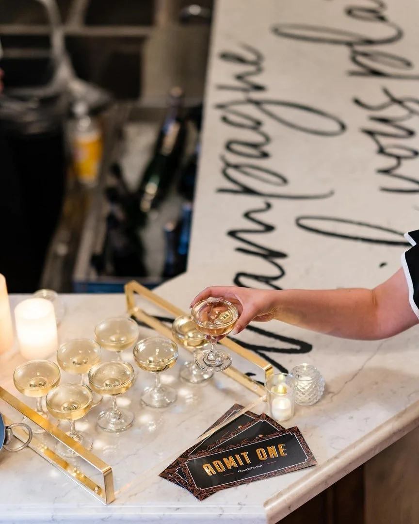A person holding a glass of champagne or sparkling wine above a tray of similar glasses on a marble countertop. The tray is gold with glass candle holders and lit candles. There are event tickets saying 'ADMIT ONE' on the counter. In the background, 