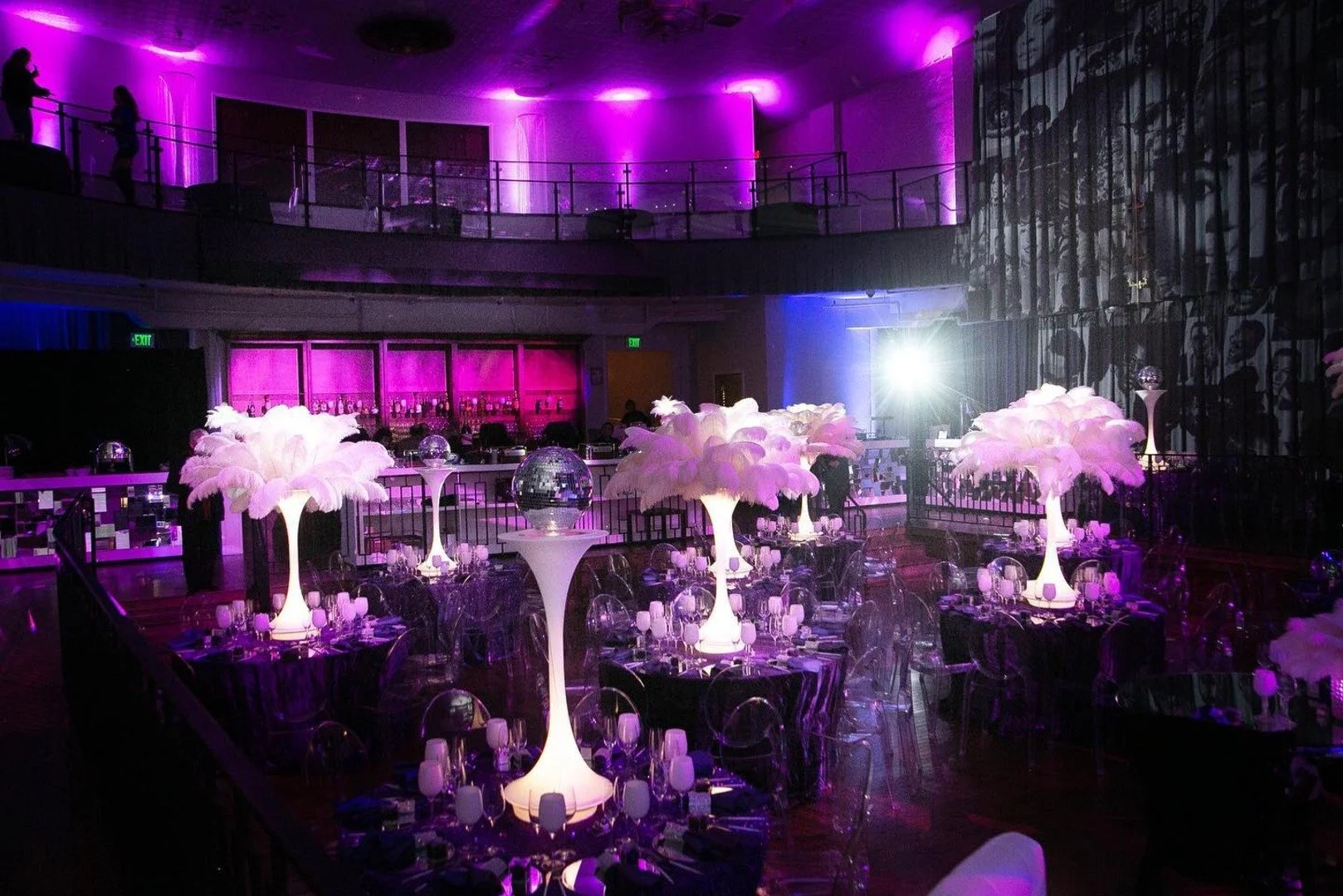 Elegant event space with round tables decorated with tall, white feathered and illuminated centerpieces, purple lighting, and a bar area in the background.