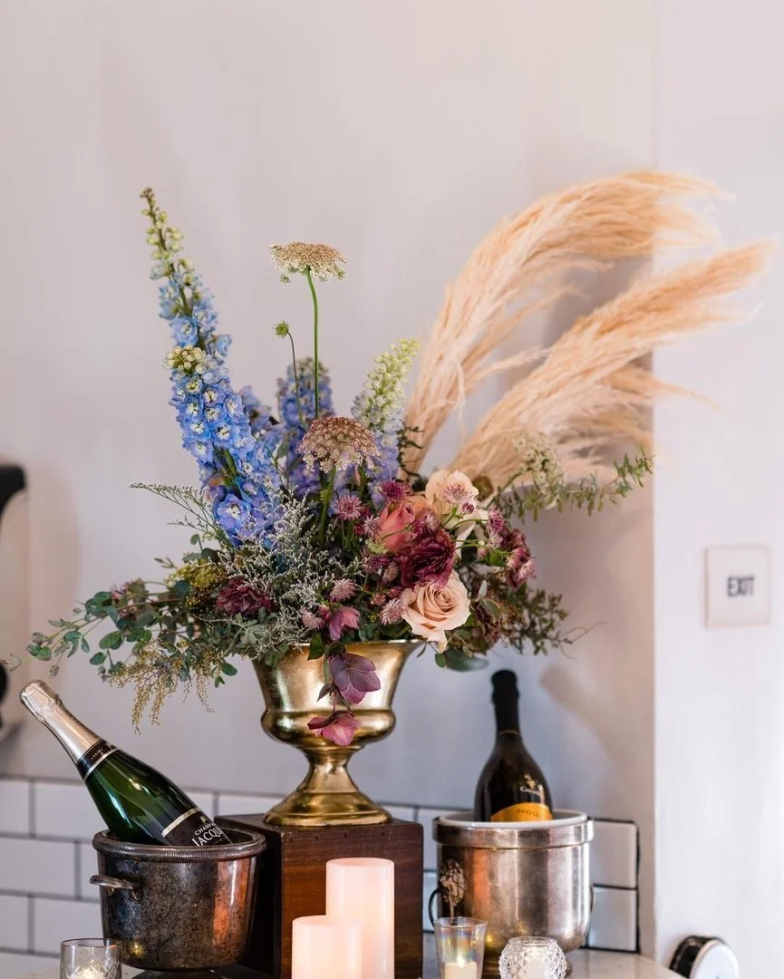 A floral arrangement with pink, purple, and blue flowers in a gold vase, with pampas grass, on a table with candles and champagne bottles