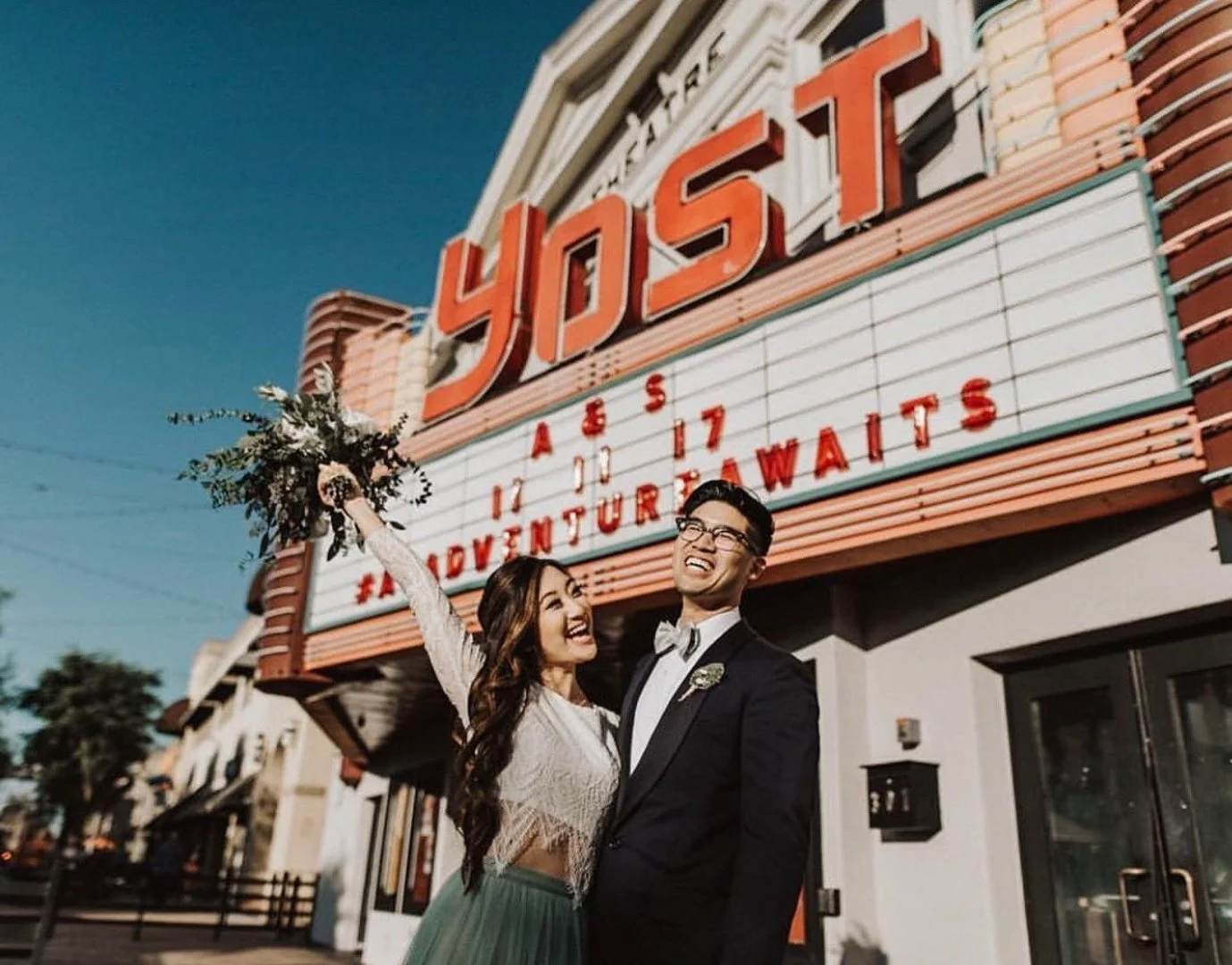A happy couple in formal attire celebrating outside a vintage Yost theater with a marquee, holding a bouquet of flowers, during the evening.