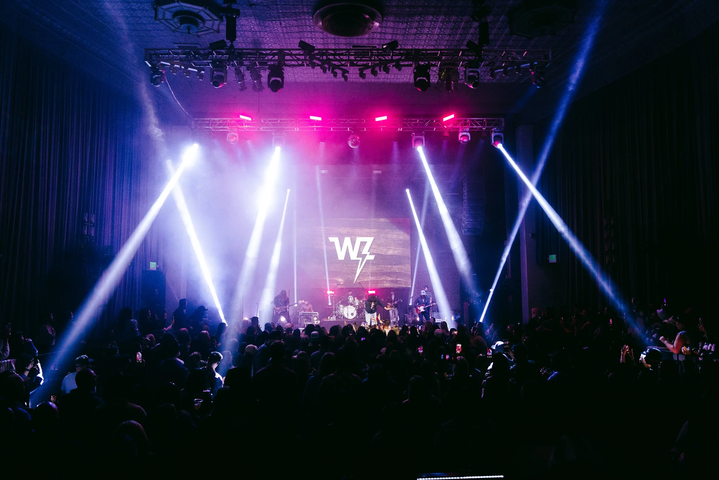 An indoor concert with a band performing on stage, colorful lights, and an audience watching.