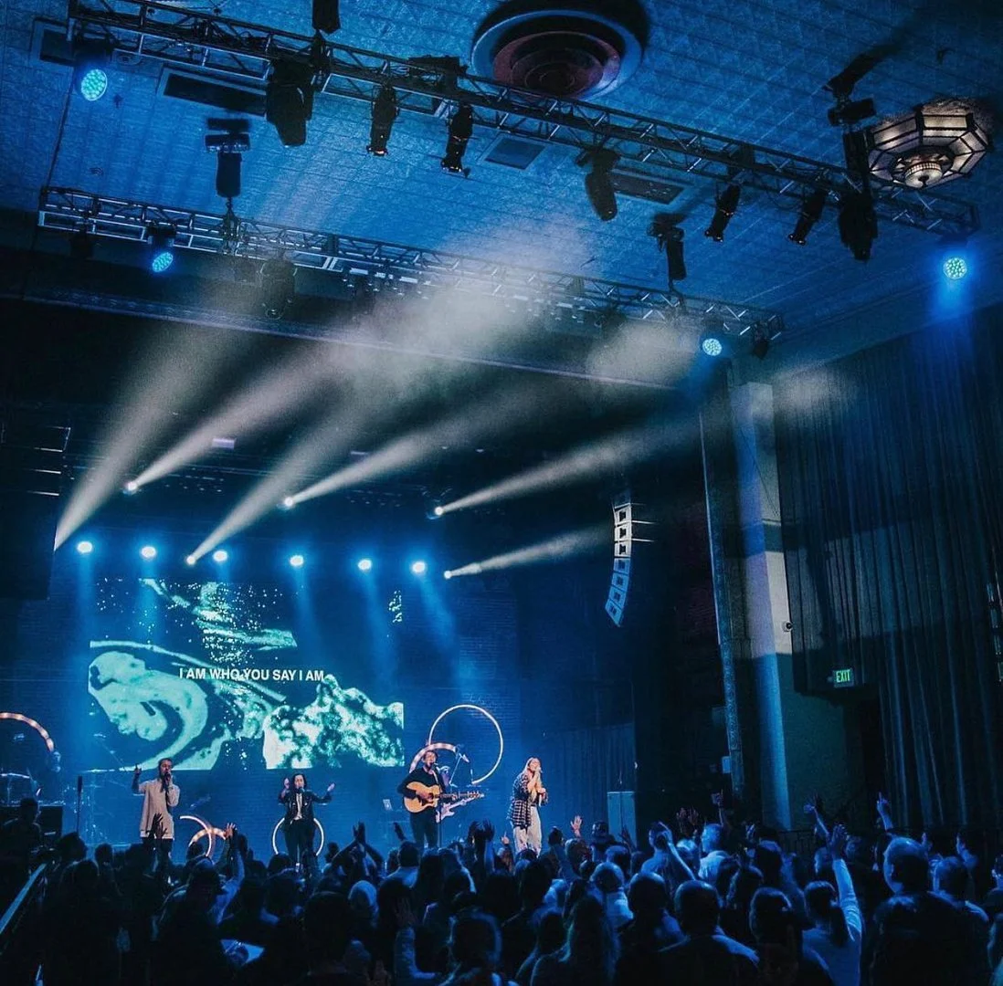 A live concert with a band performing on stage, colorful lighting, and an audience raising their hands.