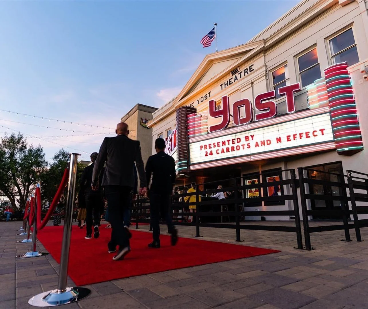 People walking on a red carpet outside a theater with a marquee that reads 'Yost,' with a sign indicating the show is presented by 24 Carrots and N Effect. The theater has a classic design with vertical neon lights and an American flag on top, and th