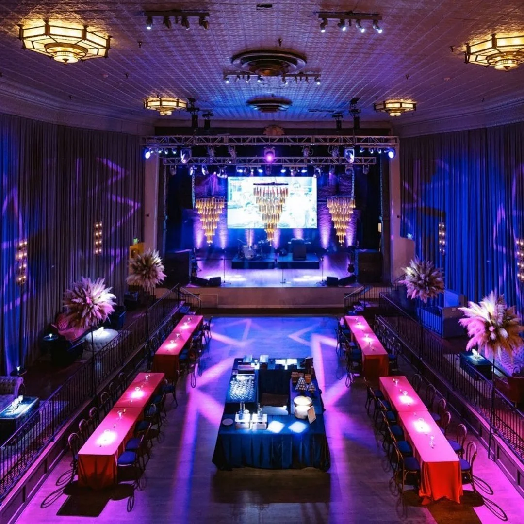 A dimly lit event space with purple and blue lighting, a stage with a large screen, floral arrangements, and long tables with red tablecloths for guests, set up for a formal event or party.