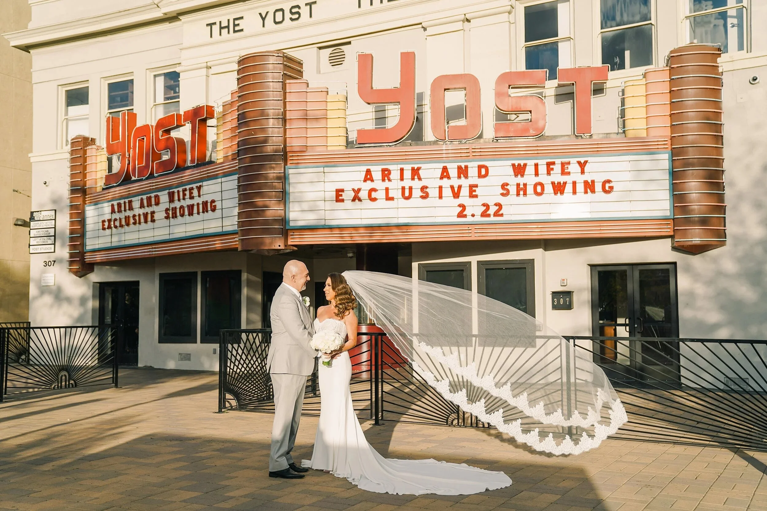 A bride and groom standing in front of a vintage theater marquee with the sign 'The Yost.' The marquee advertises an exclusive showing of Arik and Wifey on February 22. The bride wears a white wedding dress with a long train and veil, and the groom wears a light gray suit. They are holding hands and smiling at each other, with a wedding bouquet next to them.