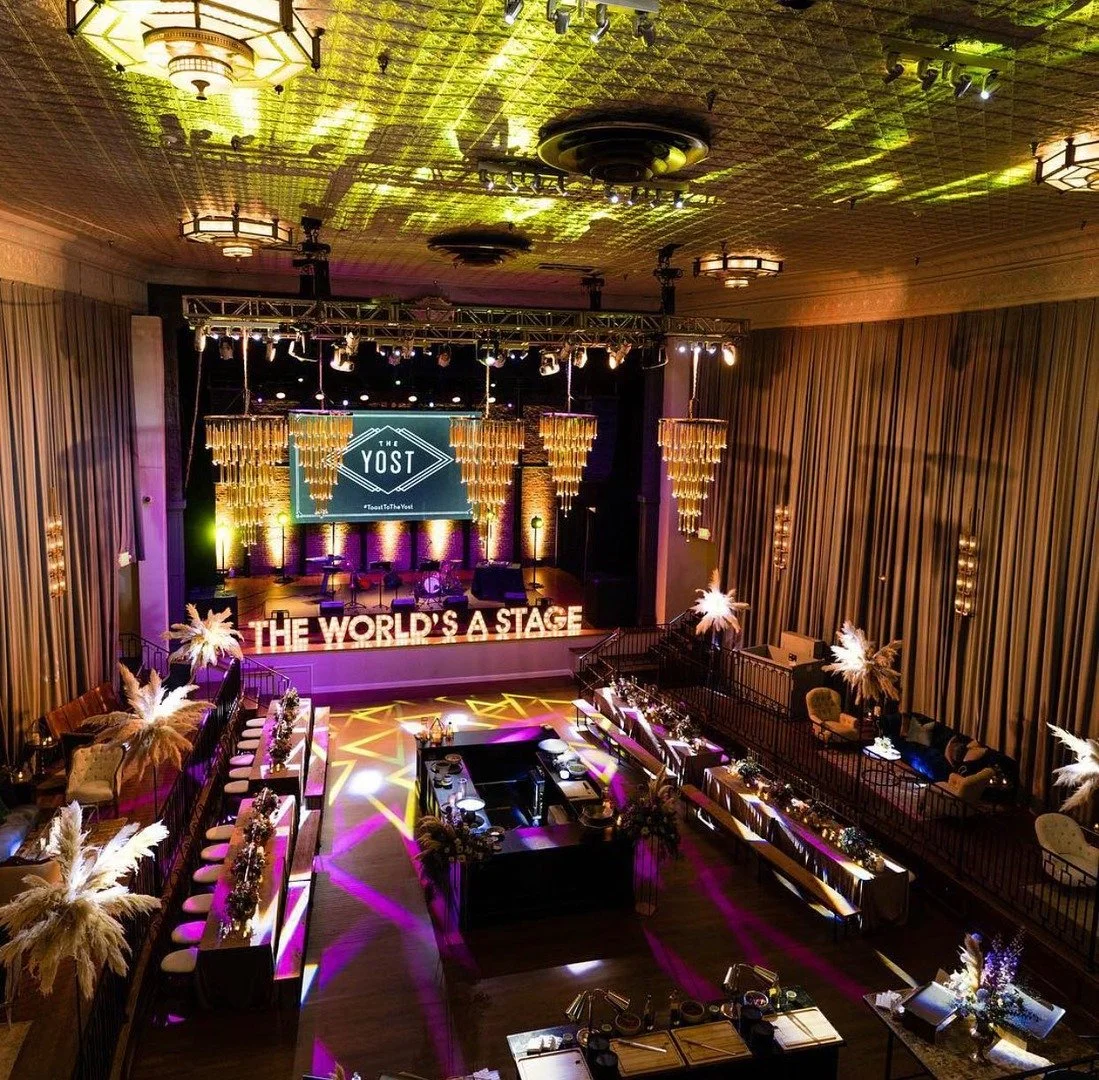 An elegant event hall with a stage decorated with hanging gold ornaments, a large screen displaying 'THE YOST,' and lights casting purple and yellow patterns on the floor. There are tables with floral arrangements, seating areas with couches and chai