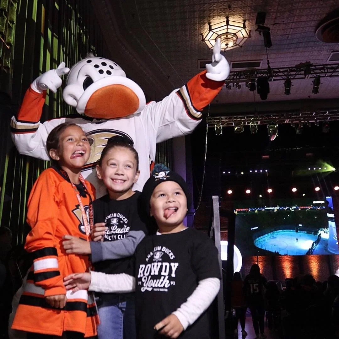 Three children posing happily with a person in a Donald Duck costume at an indoor event near an ice hockey rink, with a large screen showing the game in the background.