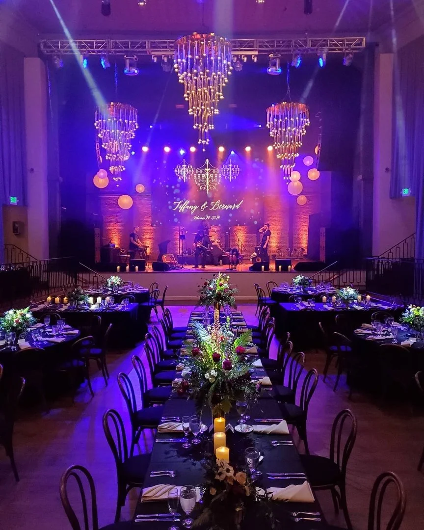 Elegant banquet hall decorated for a wedding reception, featuring a stage with band performing, chandeliers, round hanging lanterns, floral centerpieces, candles, and festively set tables