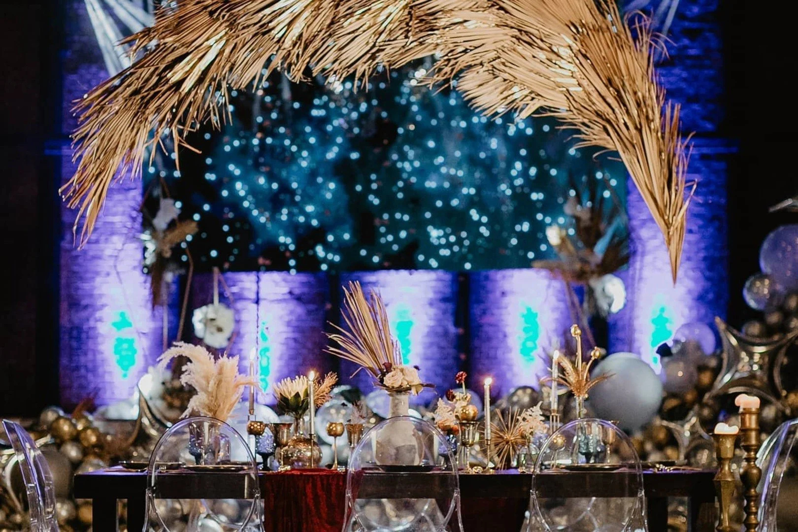 Christmas or holiday party decor featuring a table with ornaments, candles, and festive decorations, with a blurred background of blue glowing trees and a straw arch.