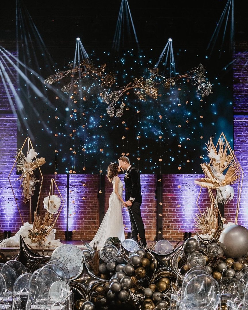 A bride and groom standing on a decorated stage during their wedding ceremony, with an elaborate backdrop of hanging floral arrangements, star-like lights, and purple uplighting on a brick wall, surrounded by metallic and clear balloons.