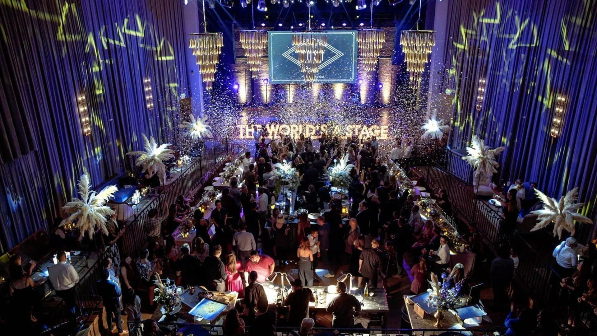 A lively party scene with many people dancing and socializing in a decorated event space, featuring large feather arrangements, hanging chandeliers, bright lighting, and a stage displaying the words 'THE WORST PLACE'.