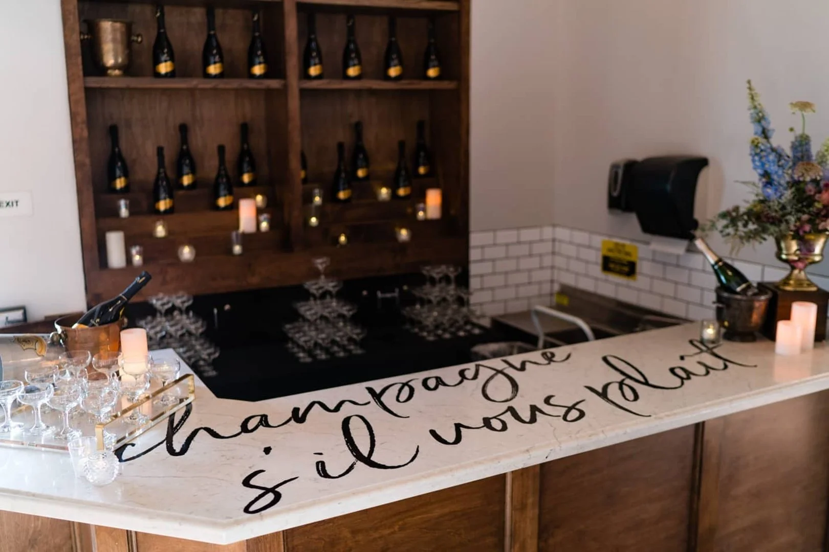 A bar counter with a white marble top featuring black cursive writing that says 'Champagne s'il vous plaît.' On the counter are champagne glasses, candles, and a champagne bucket with a bottle. In the background, a wooden shelf displays bottles of ch