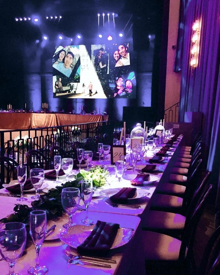 Elegant banquet table set with glasses, plates, napkins, and floral centerpiece, overlooking a stage with a large screen displaying photos of people, in a dimly lit event hall.
