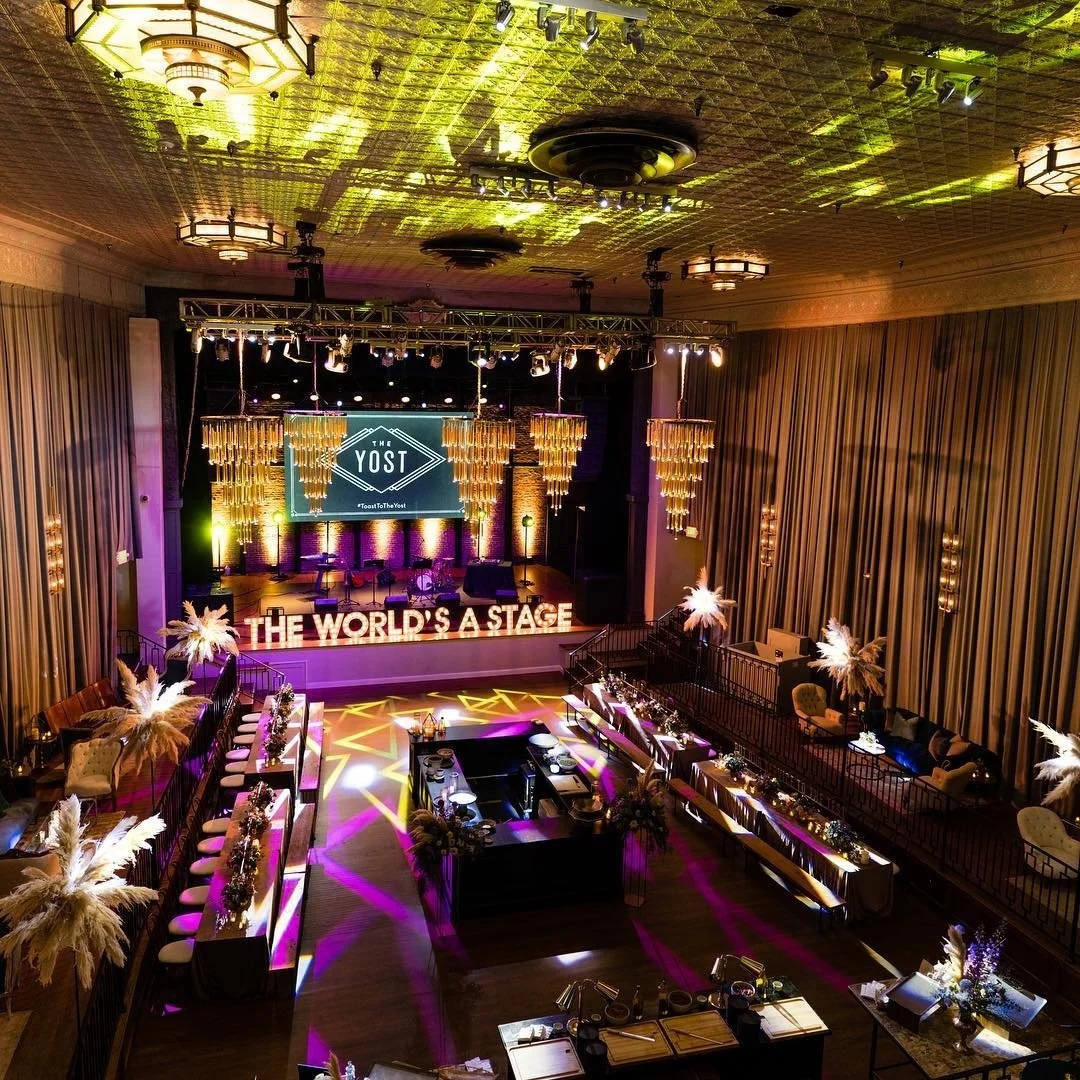 Elegant event venue with a stage featuring a large screen displaying 'THE YOST' and a hashtag. The stage is decorated with golden hanging ornaments and large illuminated letters spelling 'THE WORLD'S A STAGE.' The room has decorated tables with centerpieces, bar tables, lounge areas with armchairs, and is lit with colorful lights and spotlights creating a vibrant atmosphere.