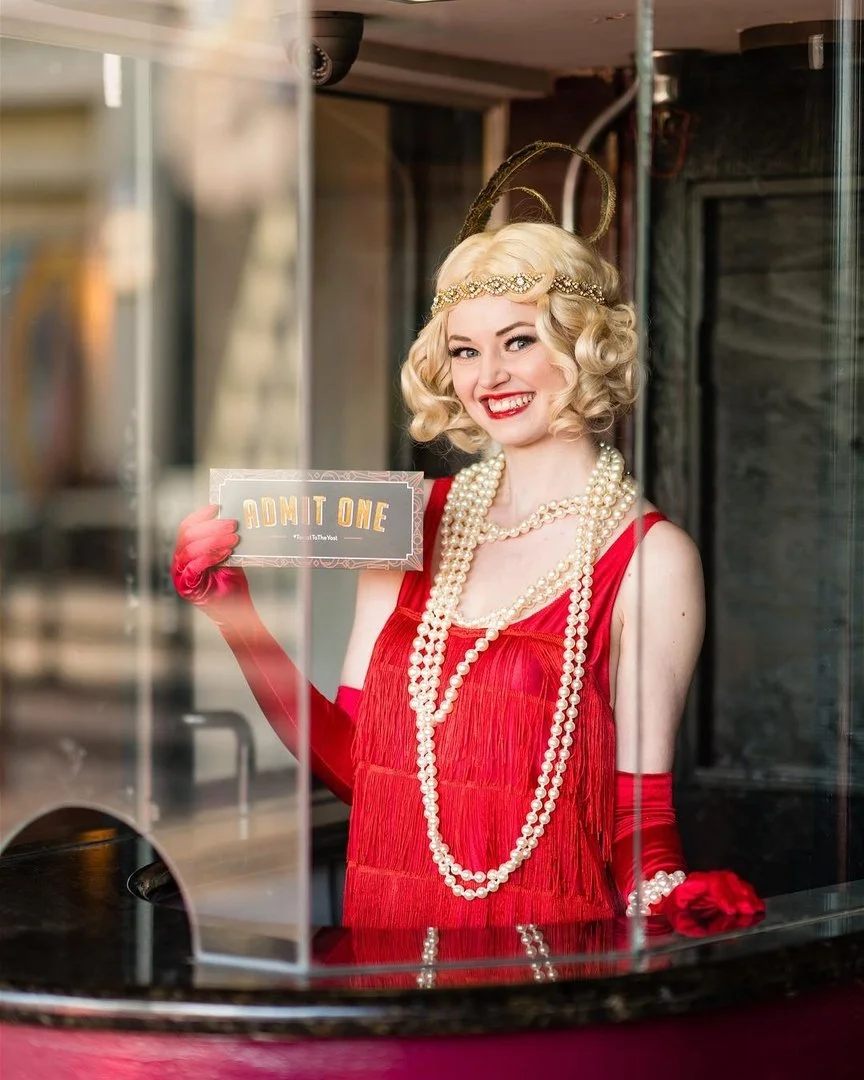 A woman dressed as a vintage showgirl with blonde curly hair, wearing a red fringe dress, multiple pearl necklaces, and red gloves, holding a ticket that reads 'ADMIT ONE', smiling through the glass window.
