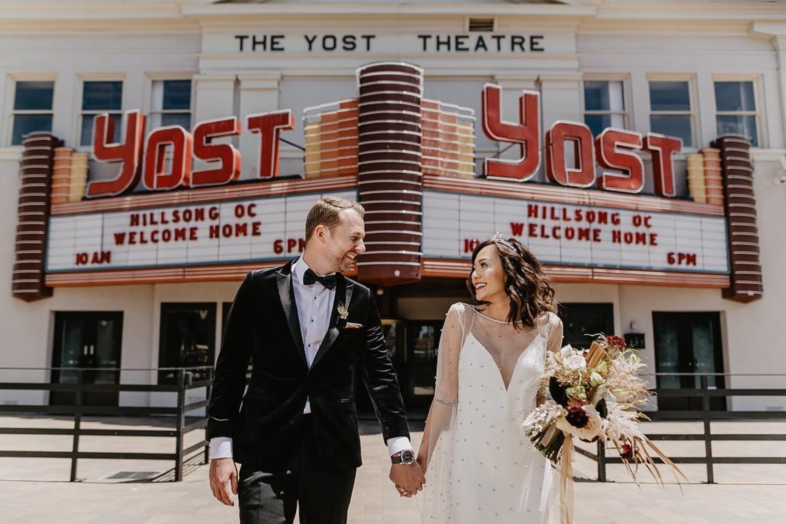 A newlywed couple holding hands and smiling in front of a vintage theater marquee, with the groom in a black tuxedo and the bride in a white wedding dress holding a bouquet of flowers.