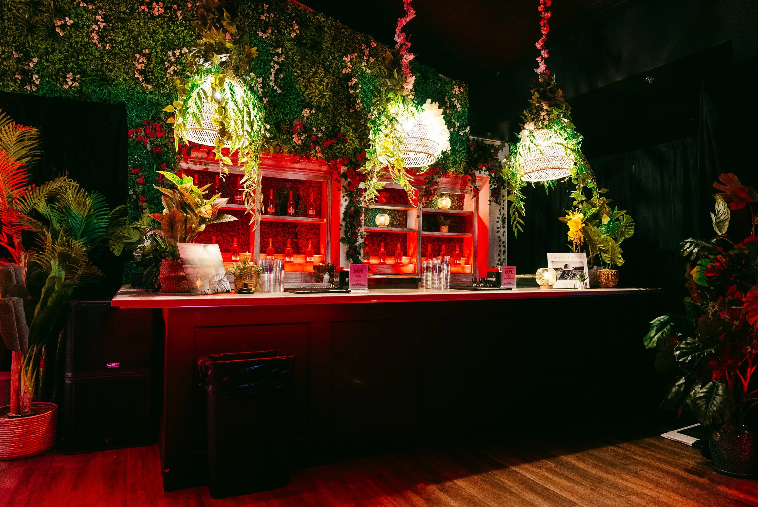A decorated bar area with green plants, pink flowers, hanging white lamps, a red backdrop, shelves with bottles, and tropical foliage in planters.