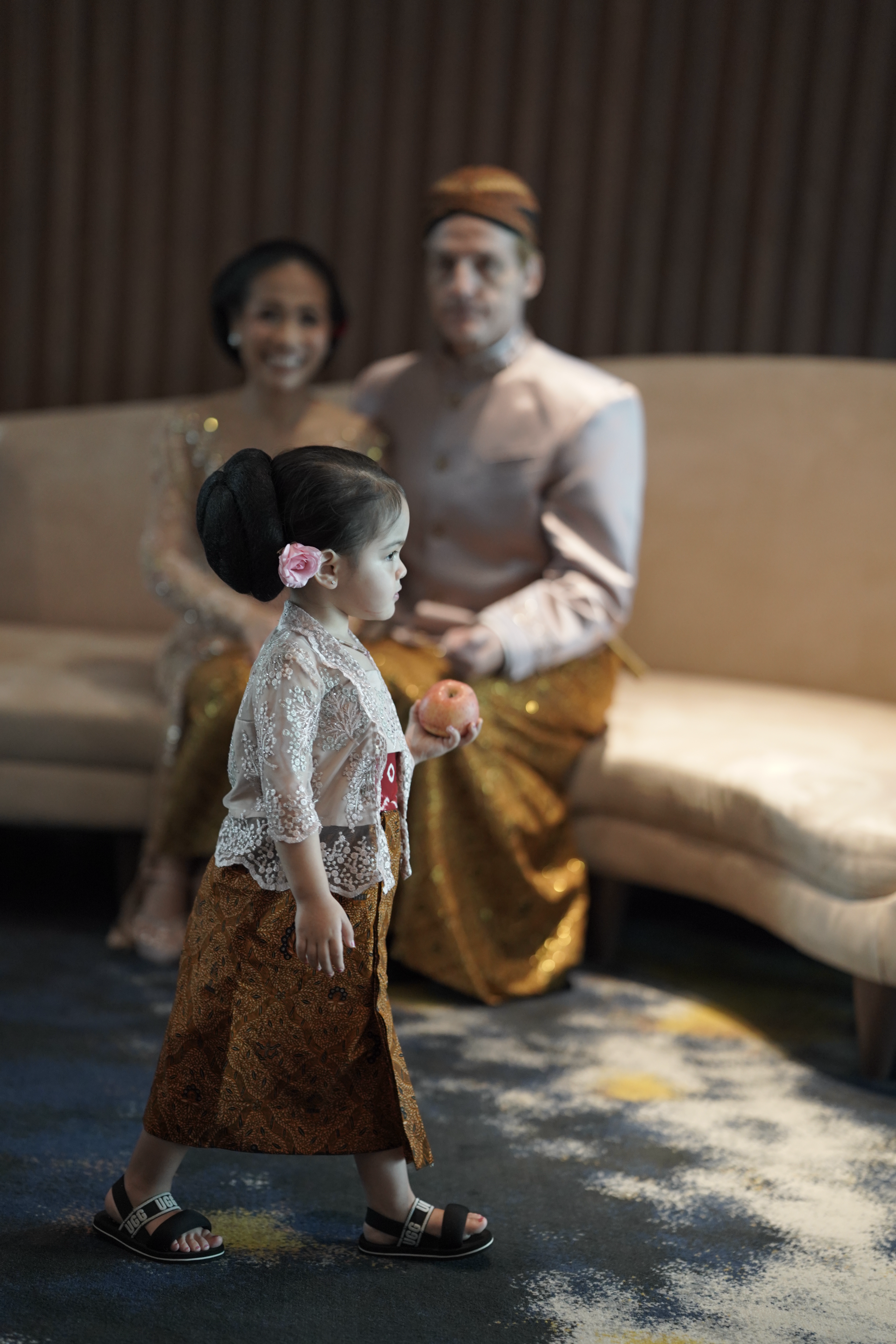 Une jeune fille en tenue traditionnelle indonésienne tenant une pomme, avec un couple en vêtements traditionnels indonésiens assis en arrière-plan.