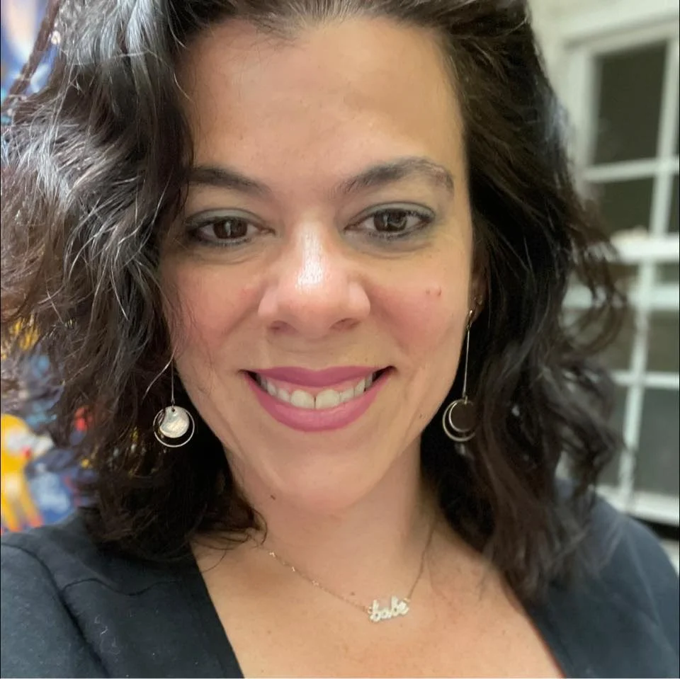 A woman with shoulder-length; curly dark hair smiling, wearing silver hoop earrings, a necklace with a nameplate, and a black top.