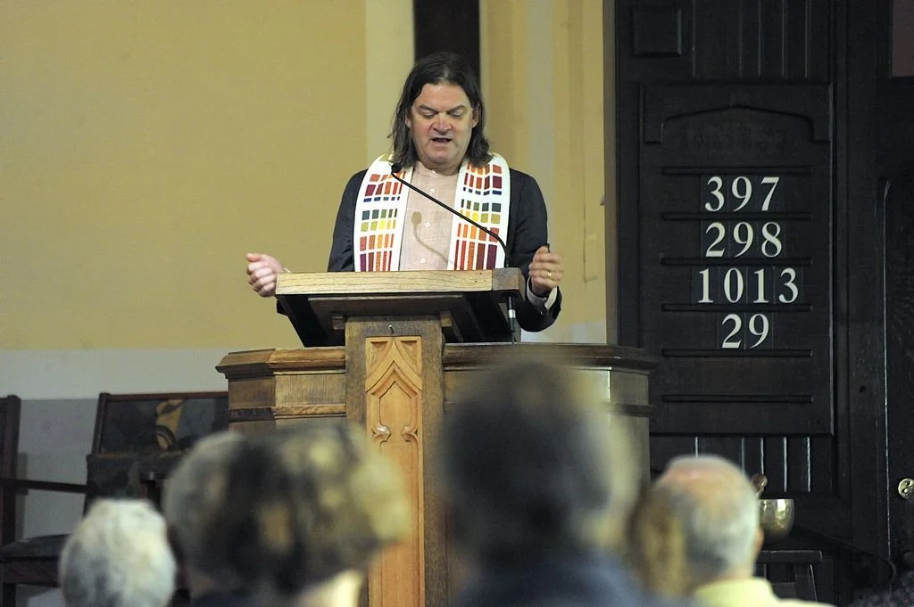 Man with long dark hair speaking at a church pulpit during a service, wearing a stole with colorful patterns.