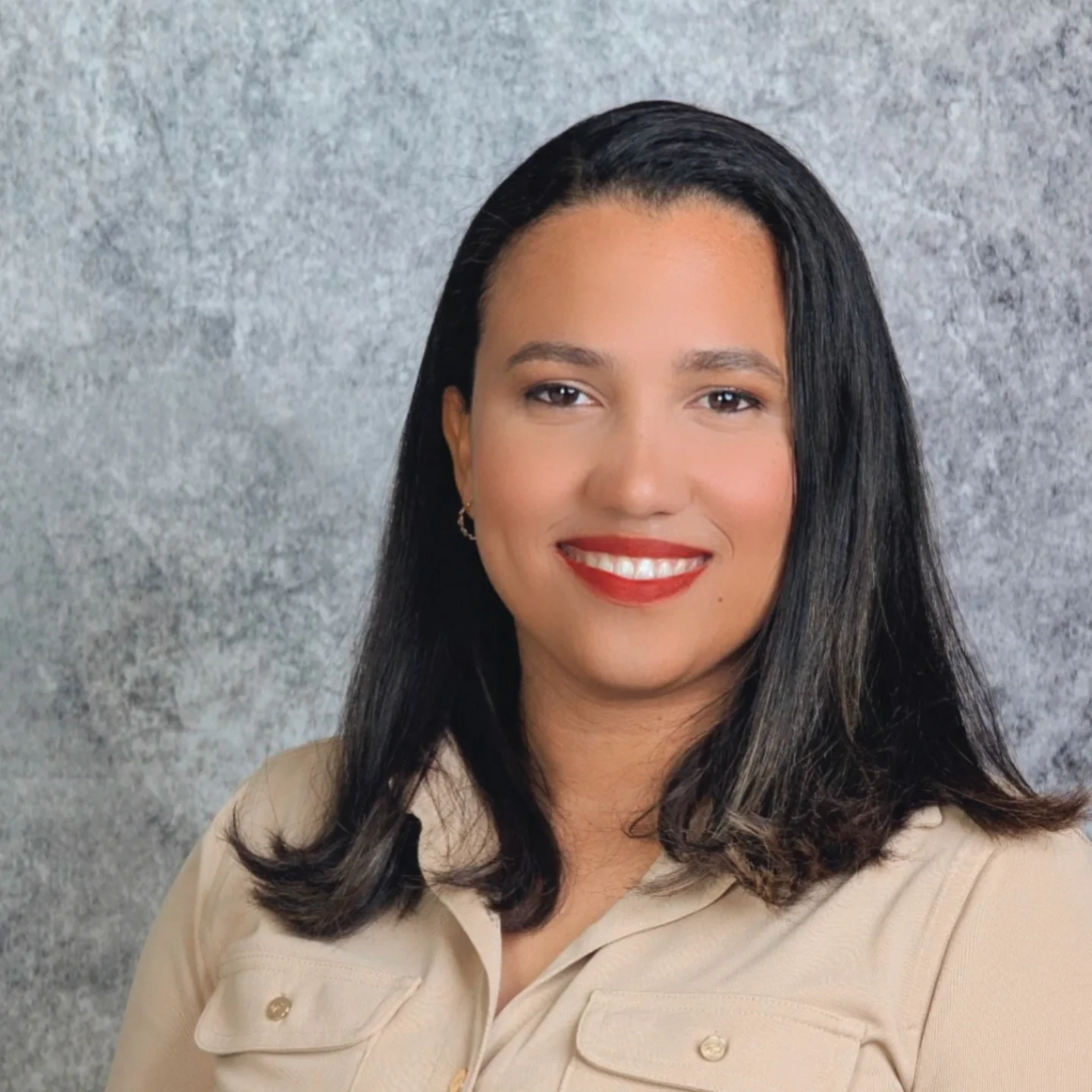 Portrait of a woman with long dark hair, smiling with red lipstick, wearing a beige collared shirt, against a textured gray background.