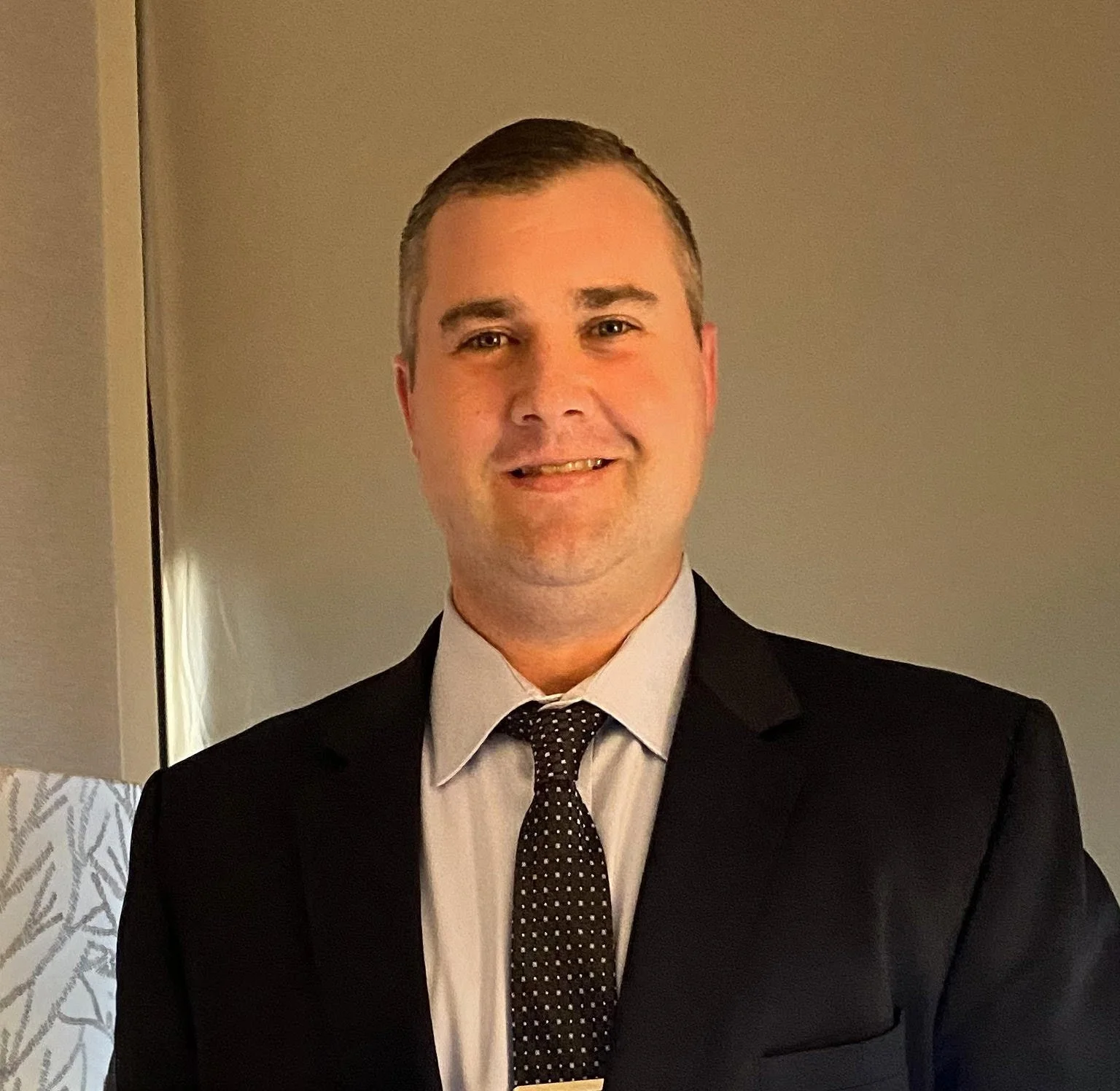 A man in a black suit with a white shirt and a dark dotted tie, smiling indoors with a plain wall in the background.