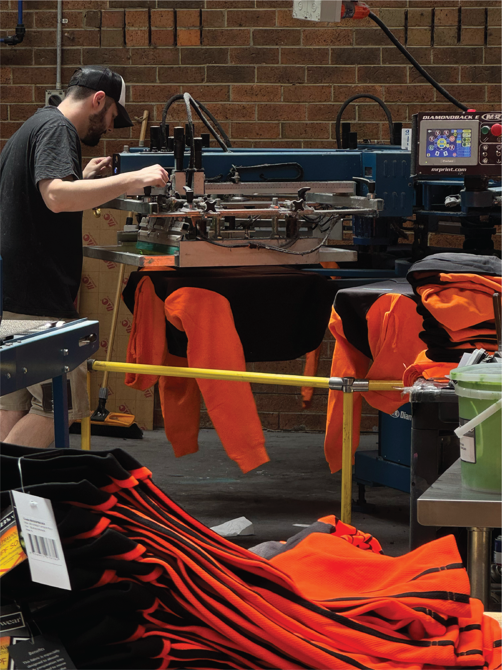 A man is working on a print manufacturing machine in a workshop, surrounded by orange and black sports apparel, with stacks of orange clothing in the foreground and a brick wall in the background.