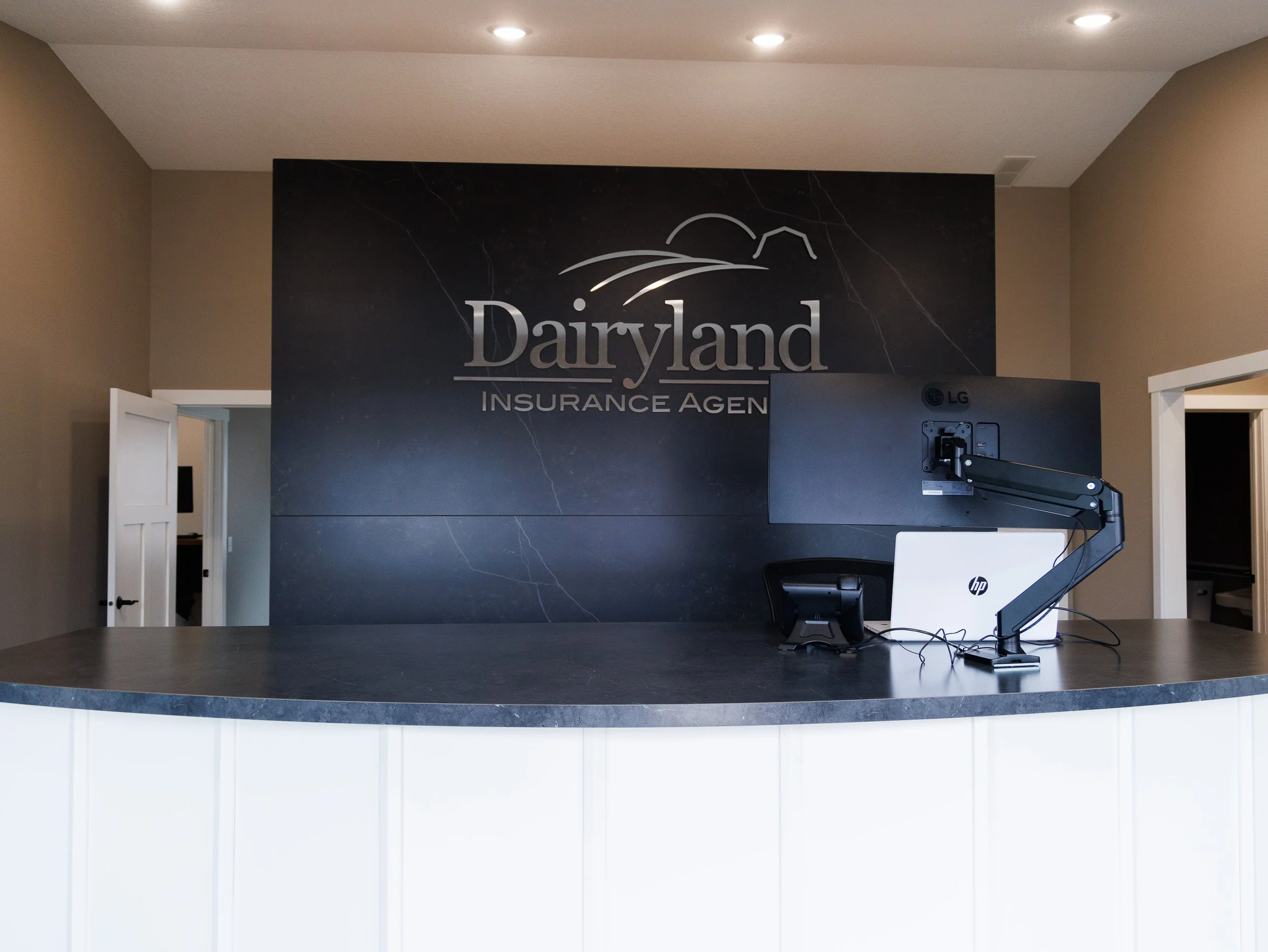 Commercial office reception area featuring a sit-stand workstation with dual monitor arms set against a dark, veined stone feature wall displaying the Dairyland Insurance Agency logo