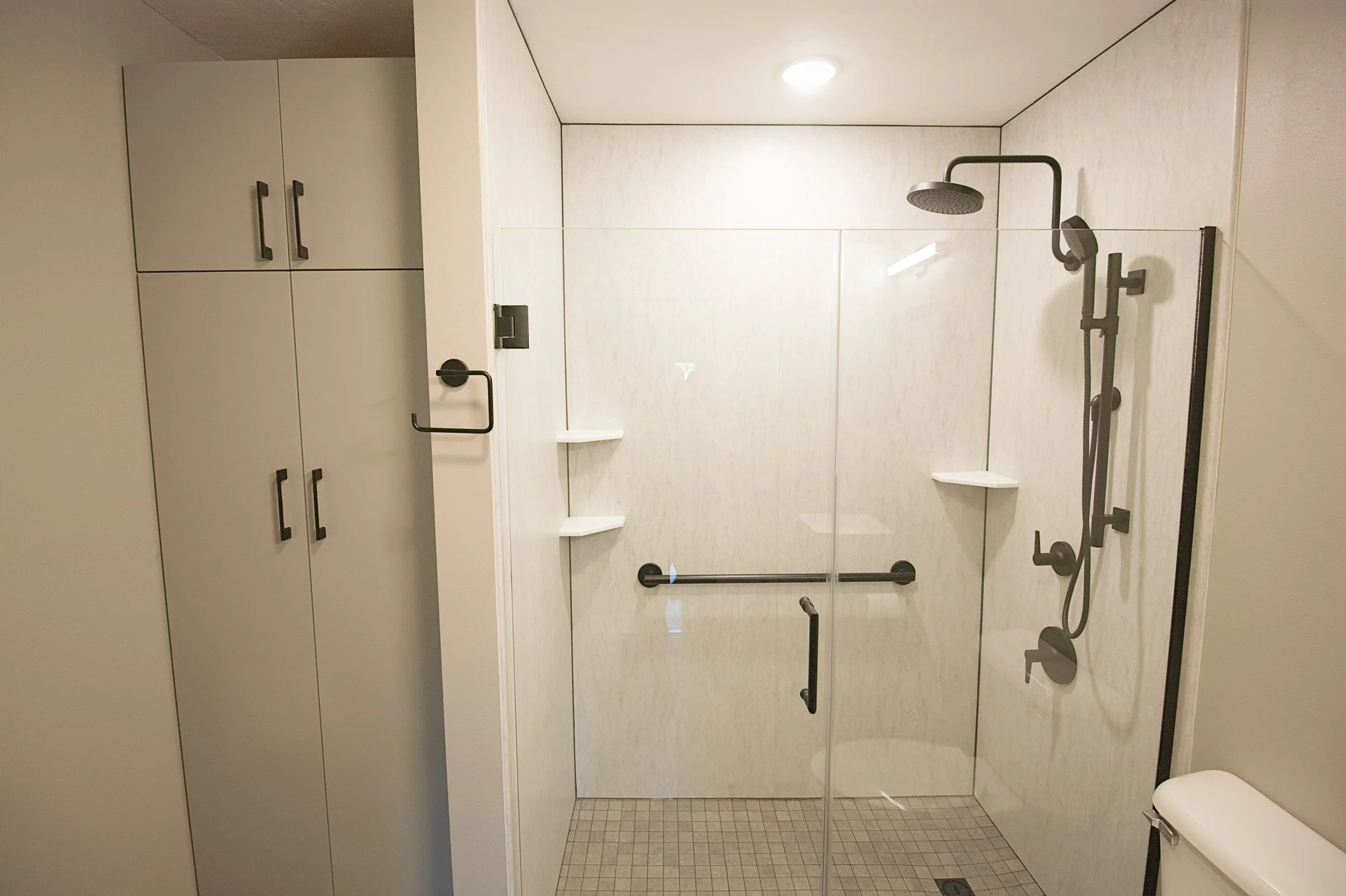 Modern bathroom remodel featuring a light greige floor-to-ceiling custom linen cabinet alongside a walk-in Wetwall shower panel surround with matte black fixtures and a frameless glass enclosure.