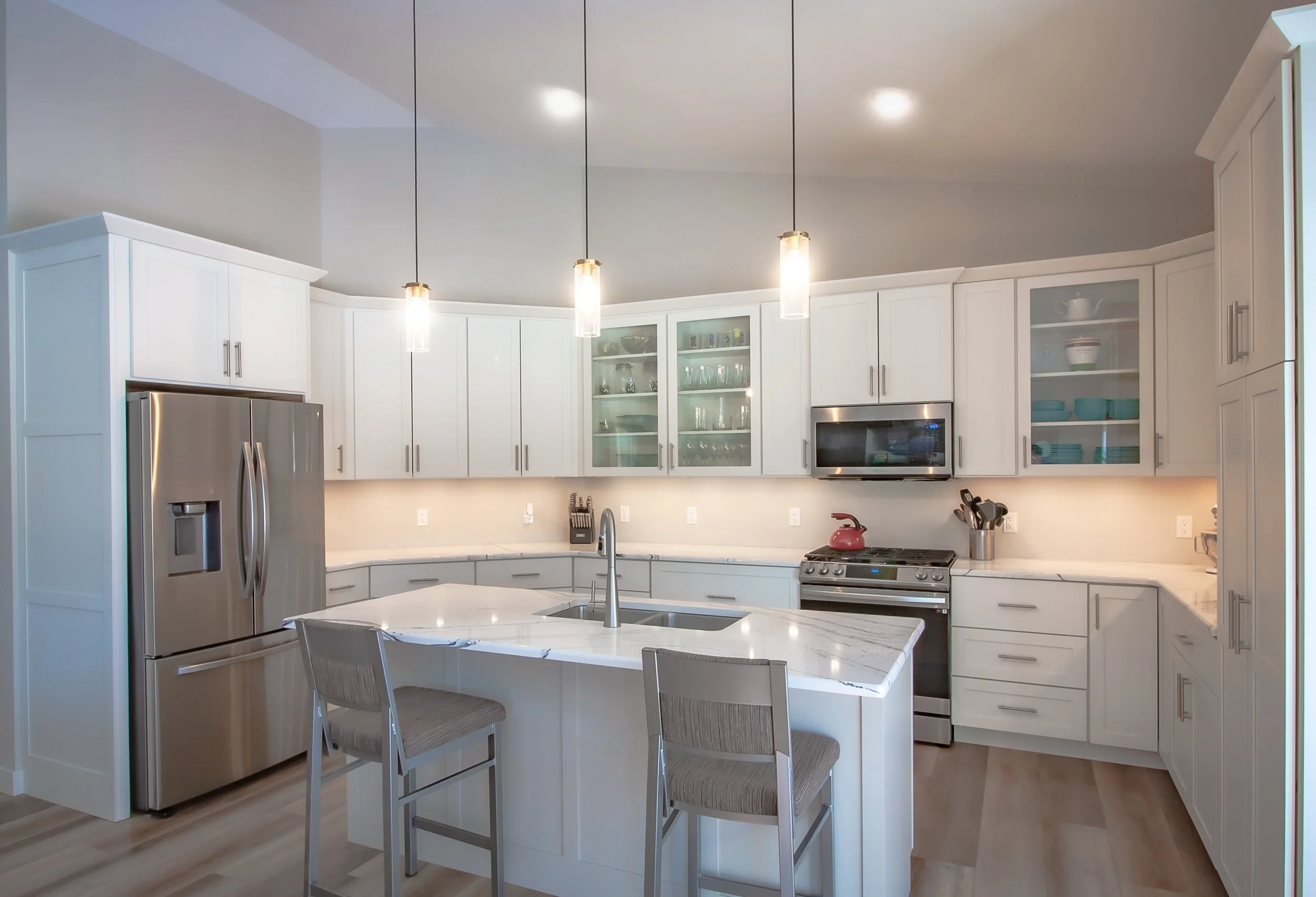 Custom white and light gray kitchen cabinetry designed to fit angled perimeter walls and a vaulted ceiling, featuring glass-front uppers, built-in refrigerator enclosure, under-cabinet lighting, and hanging pendant lights.