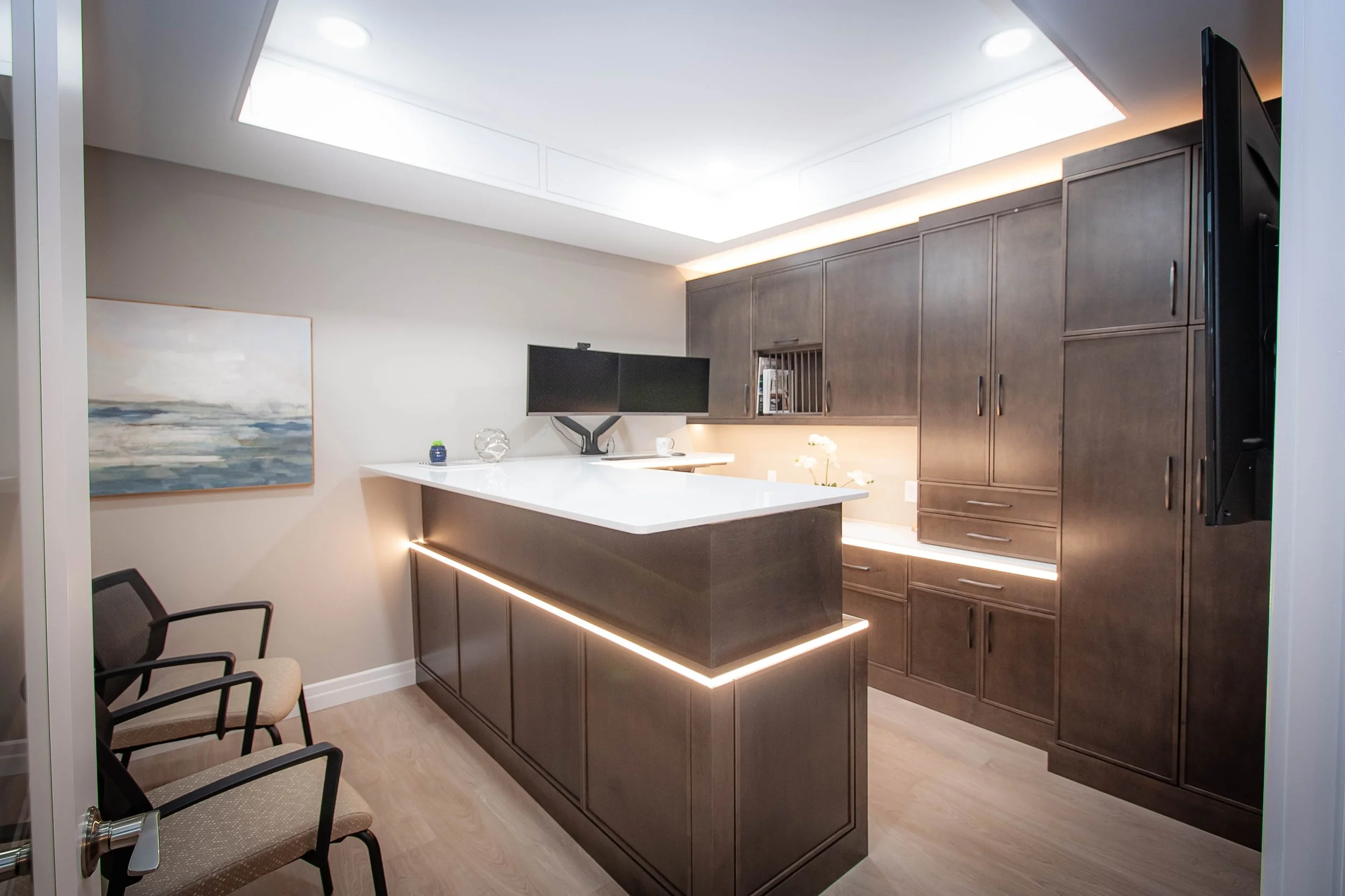 Modern commercial reception area featuring an adjustable-height standing desk with LED strip lighting, dual monitors, and custom dark wood cabinetry with an illuminated ceiling panel.