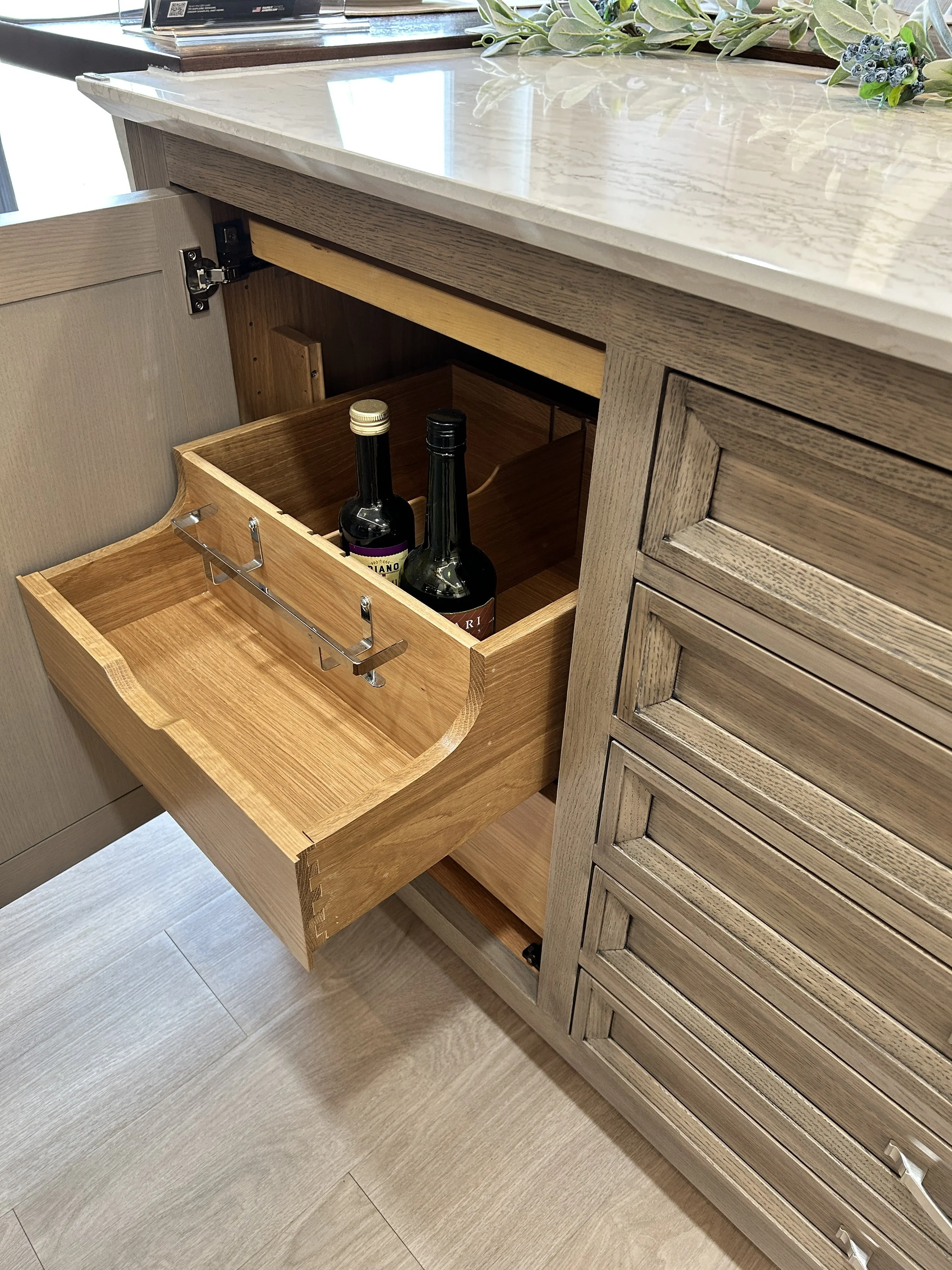 Custom light wood corner cabinet display featuring a smooth pull-out storage drawer mechanism holding bottles inside the Crown Cabinetry showroom.