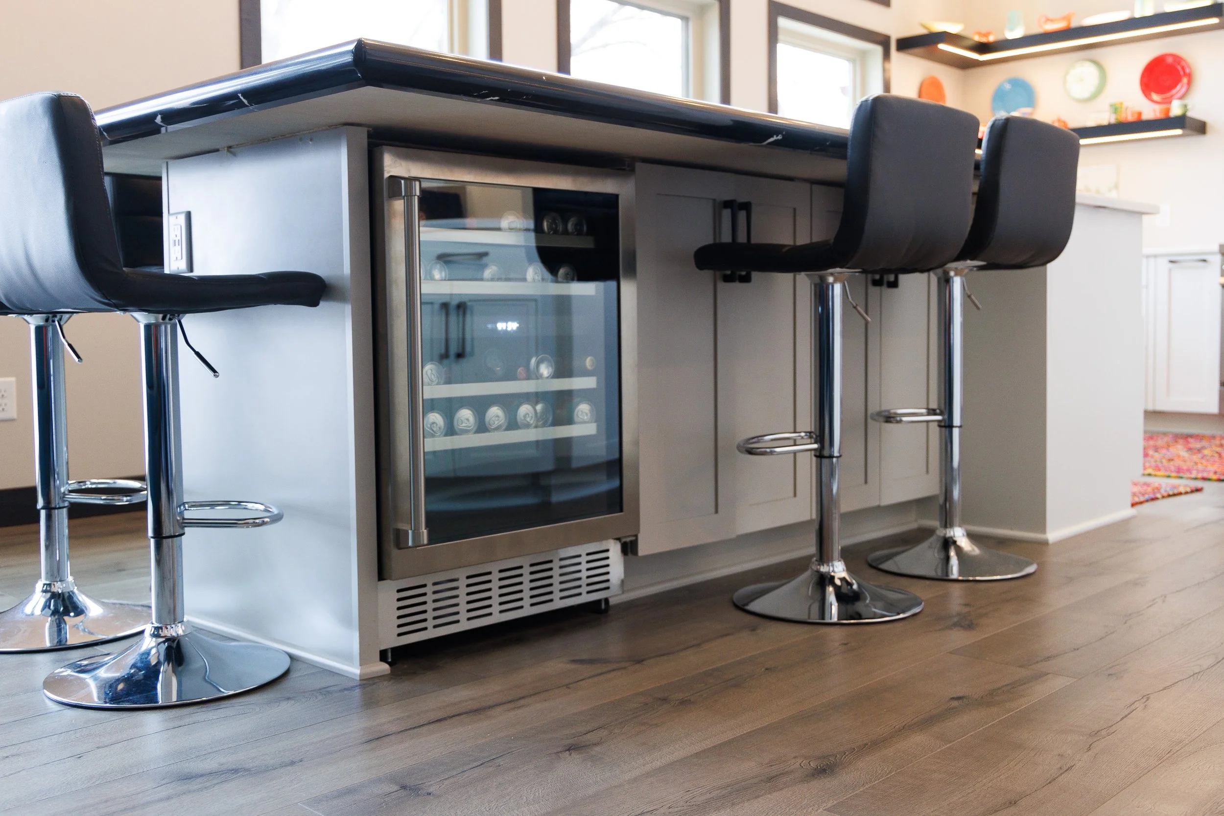 Custom kitchen island featuring an integrated stainless steel under-counter beverage cooler, light gray shaker cabinets, side electrical outlet, and modern bar stools.