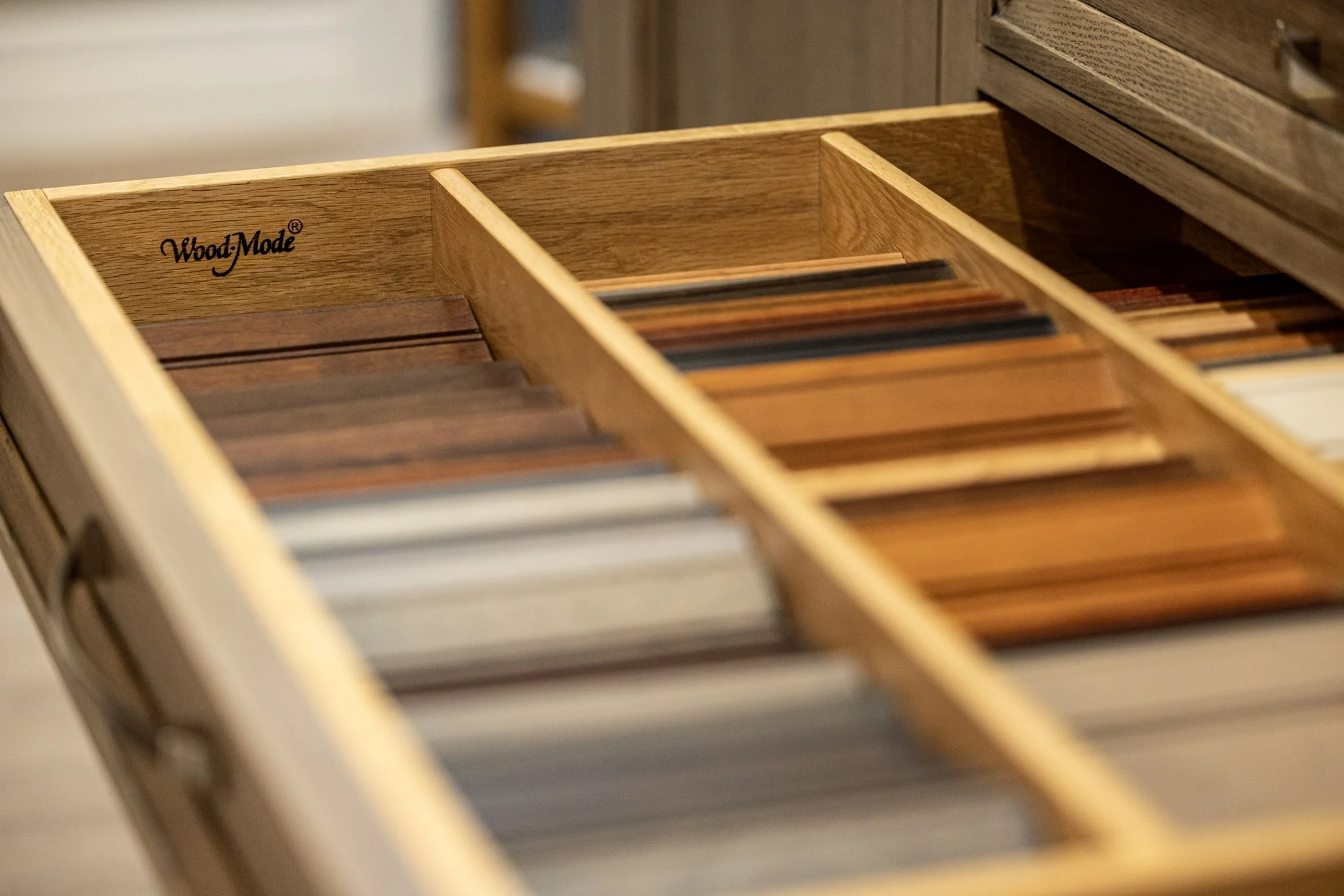 A wooden drawer with compartments, brand labeled 'WoodMode', containing wooden samples or veneers, inside a woodworking or furniture shop.