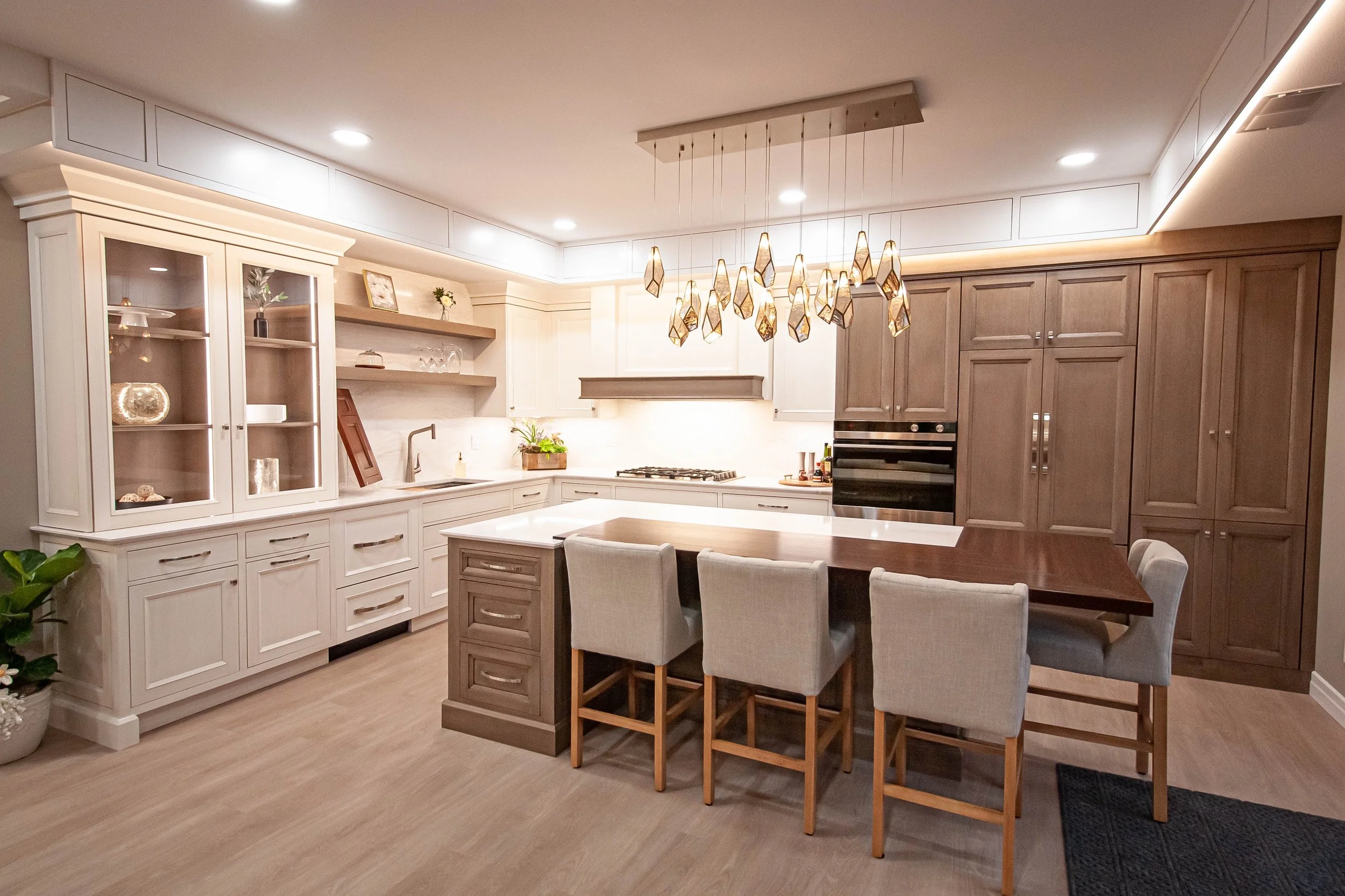 Modern custom kitchen showroom display featuring a large center island with seating, ambient pendant lighting, and warm wood and painted cabinetry.