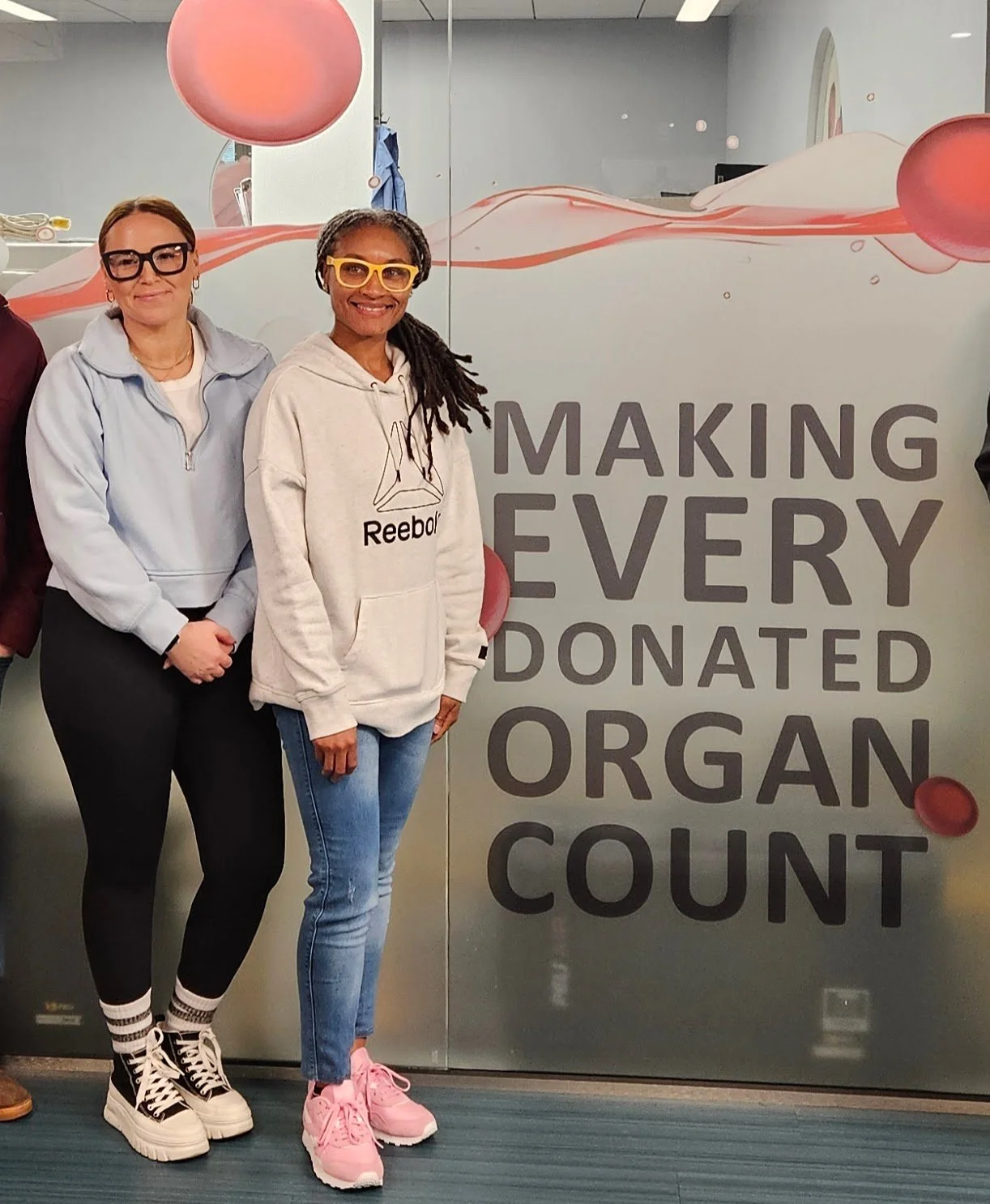 Two women standing in front of a glass wall with the text 'Making Every Donated Organ Count' and pink decorative elements. One woman wears glasses, a light gray Reebok hoodie, and pink sneakers. The other woman wears glasses, a white jacket, black pants, and black-and-white sneakers.