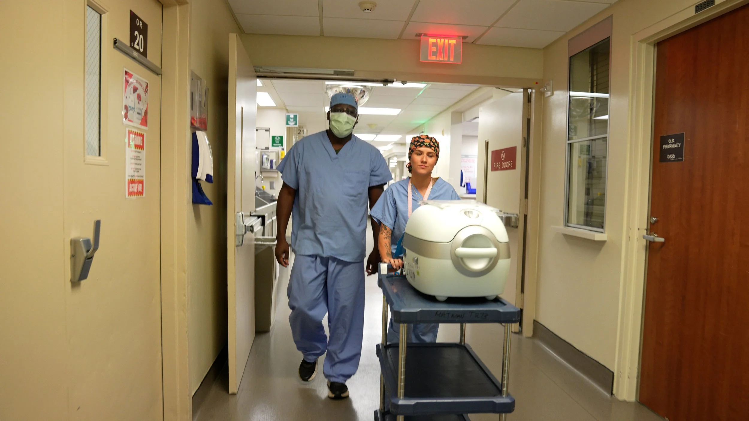 Two healthcare professionals, a male and a female, dressed in scrubs and face masks, walking down a hospital corridor with medical equipment and signs in the background.