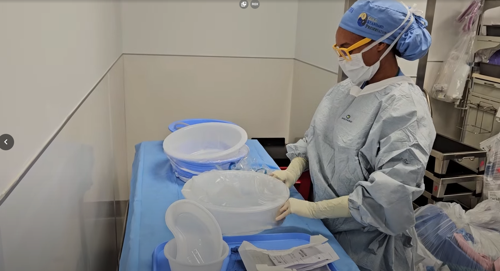 Healthcare worker wearing protective gear and a mask preparing sterile containers on a blue covered table in a medical setting.