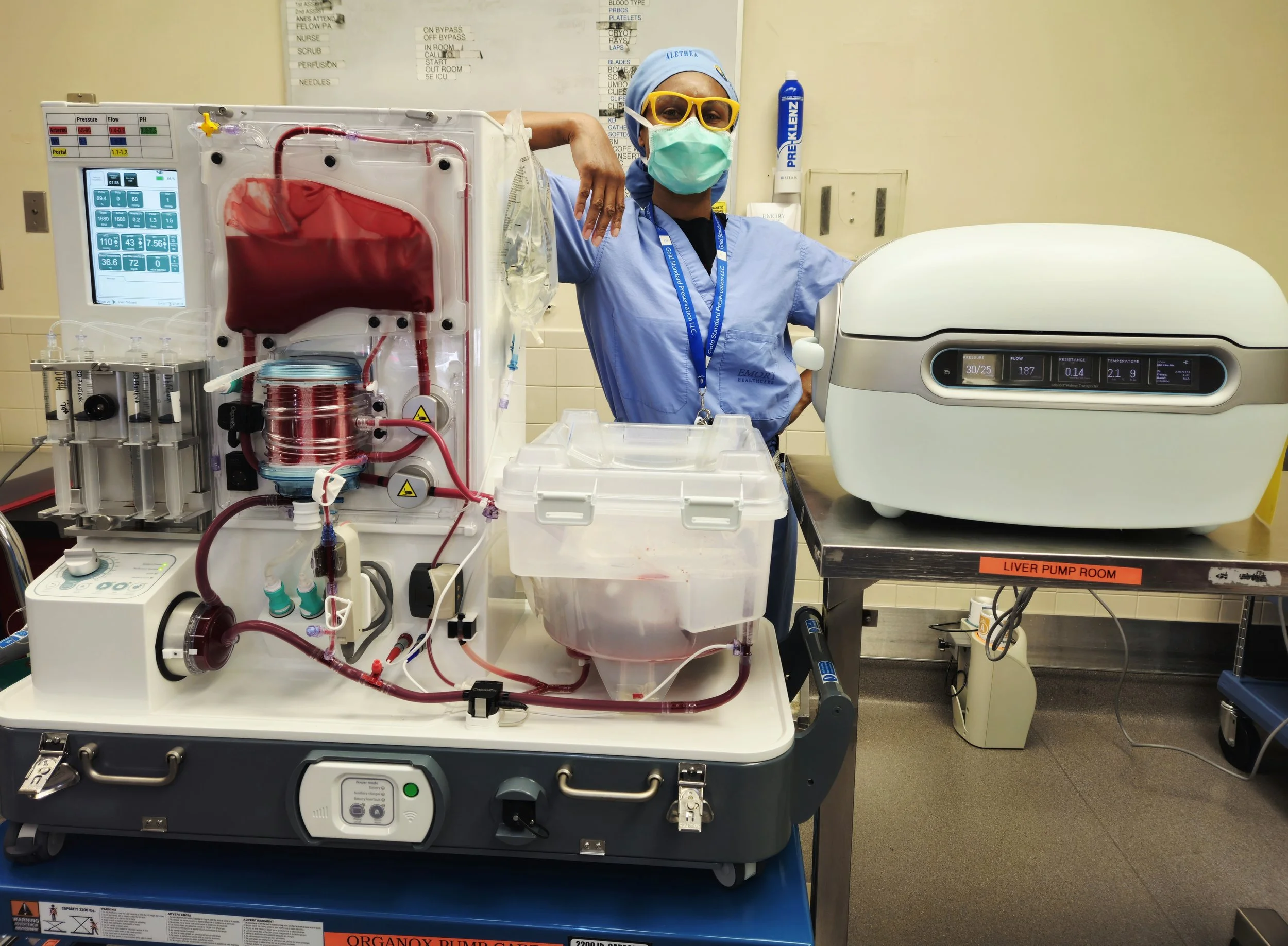 A healthcare professional in scrubs, wearing a face mask, goggles, and a hair cover, stands next to a medical device with tubes and a blood-filled chamber, in a hospital room labeled 'Liver Pump Room'.