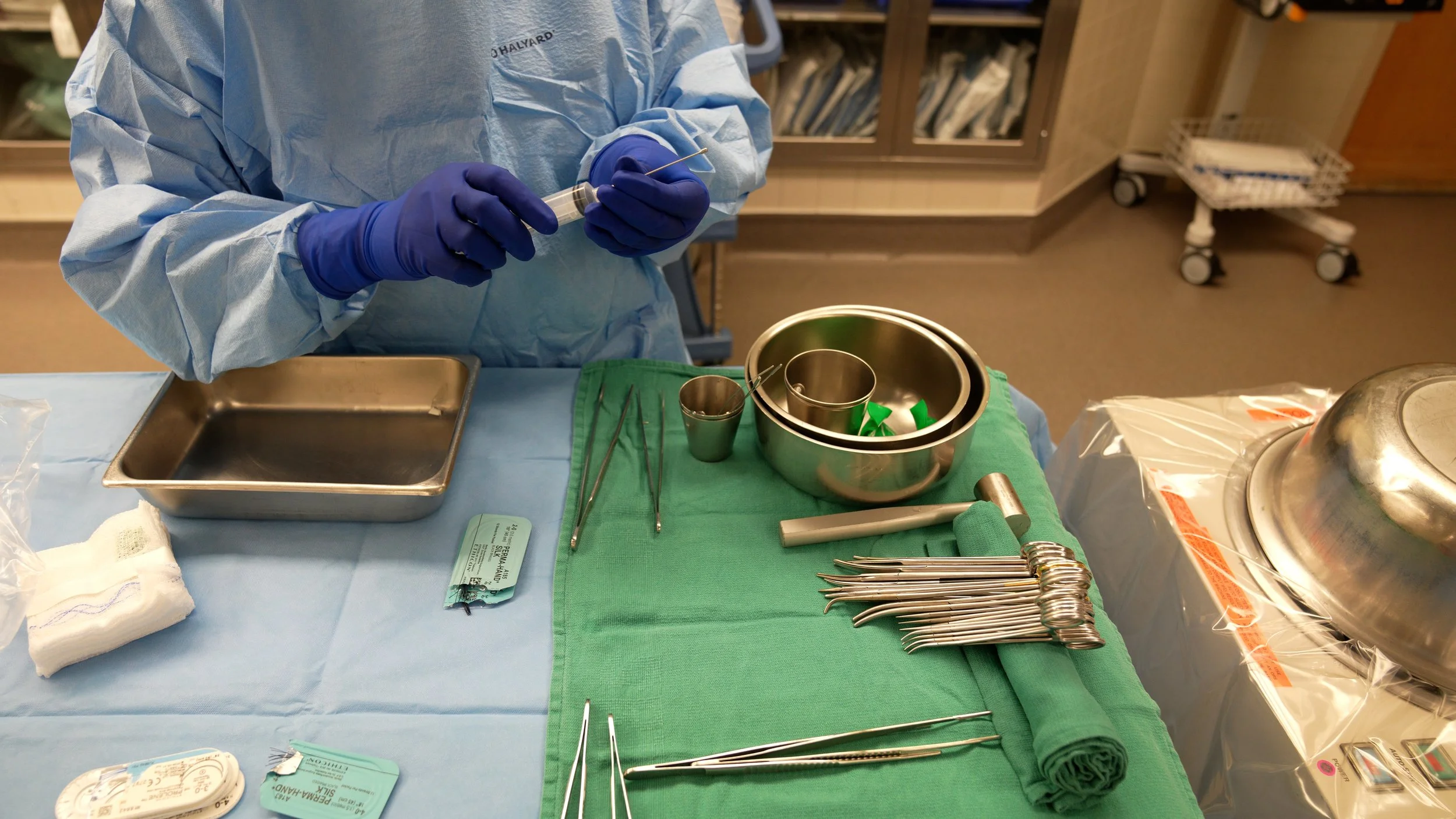 Medical professional preparing surgical instruments on a hospital tray, with various metal tools, a green cloth, and a blue gown.