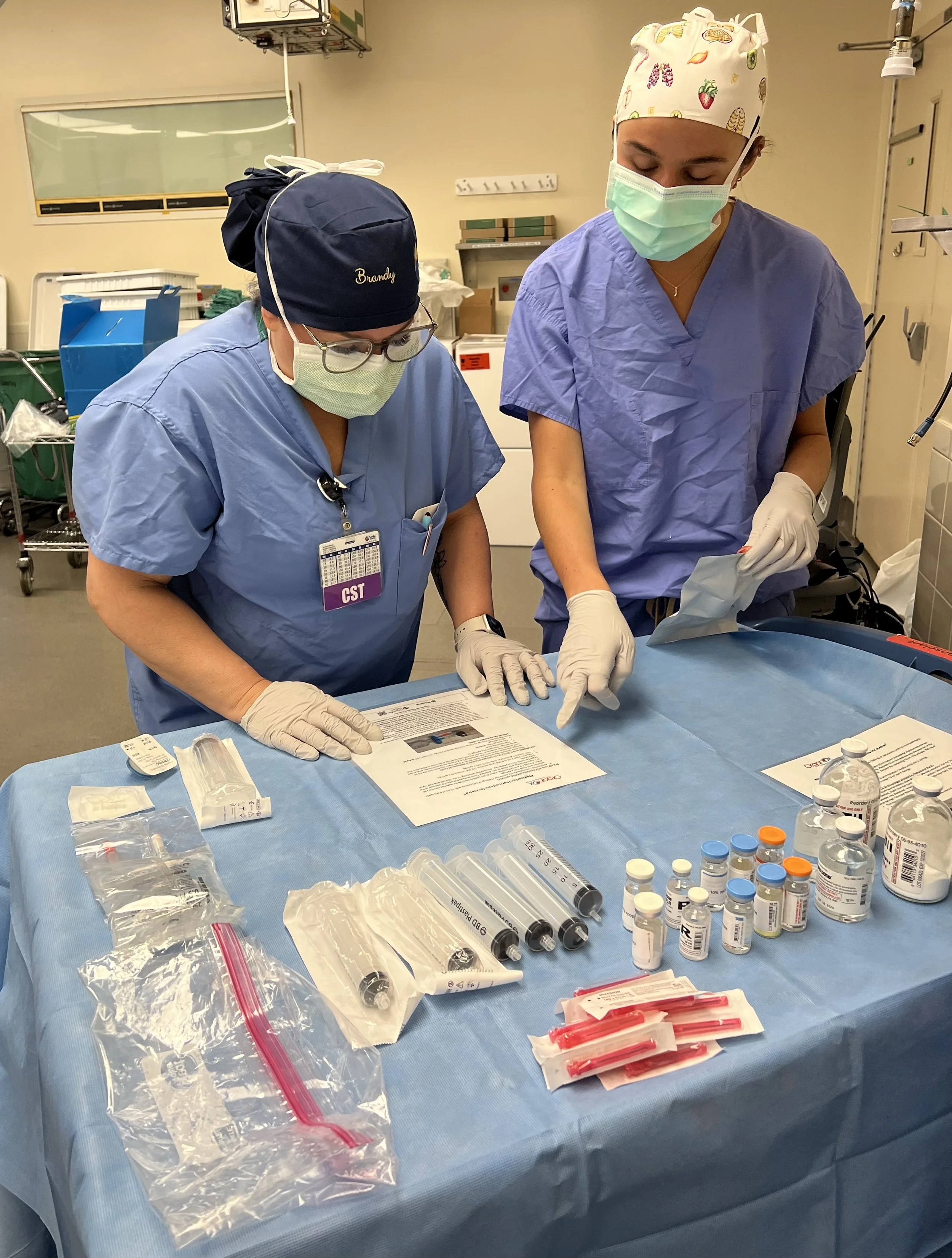 Two healthcare professionals wearing masks, gloves, and scrubs working at a table with medical supplies in a clinical setting.