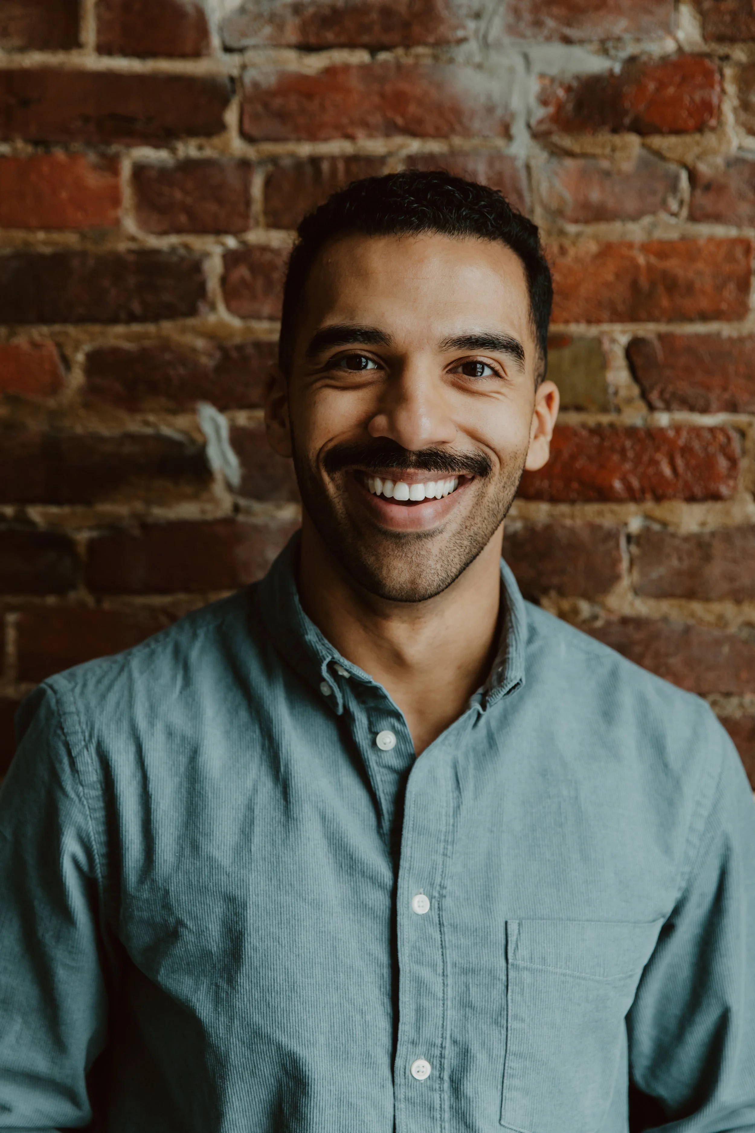 A smiling man with dark hair, a mustache, and beard, wearing a light blue button-up shirt, standing in front of a red brick wall.