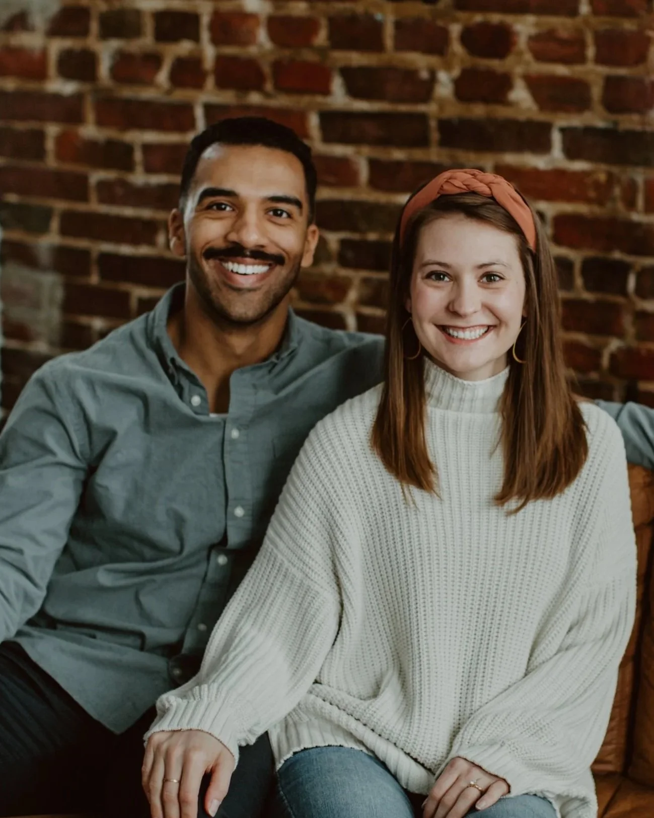 A smiling man and woman sitting together on a couch in front of a brick wall, with the man wearing a grey button-up shirt and the woman wearing a white sweater and a headband.