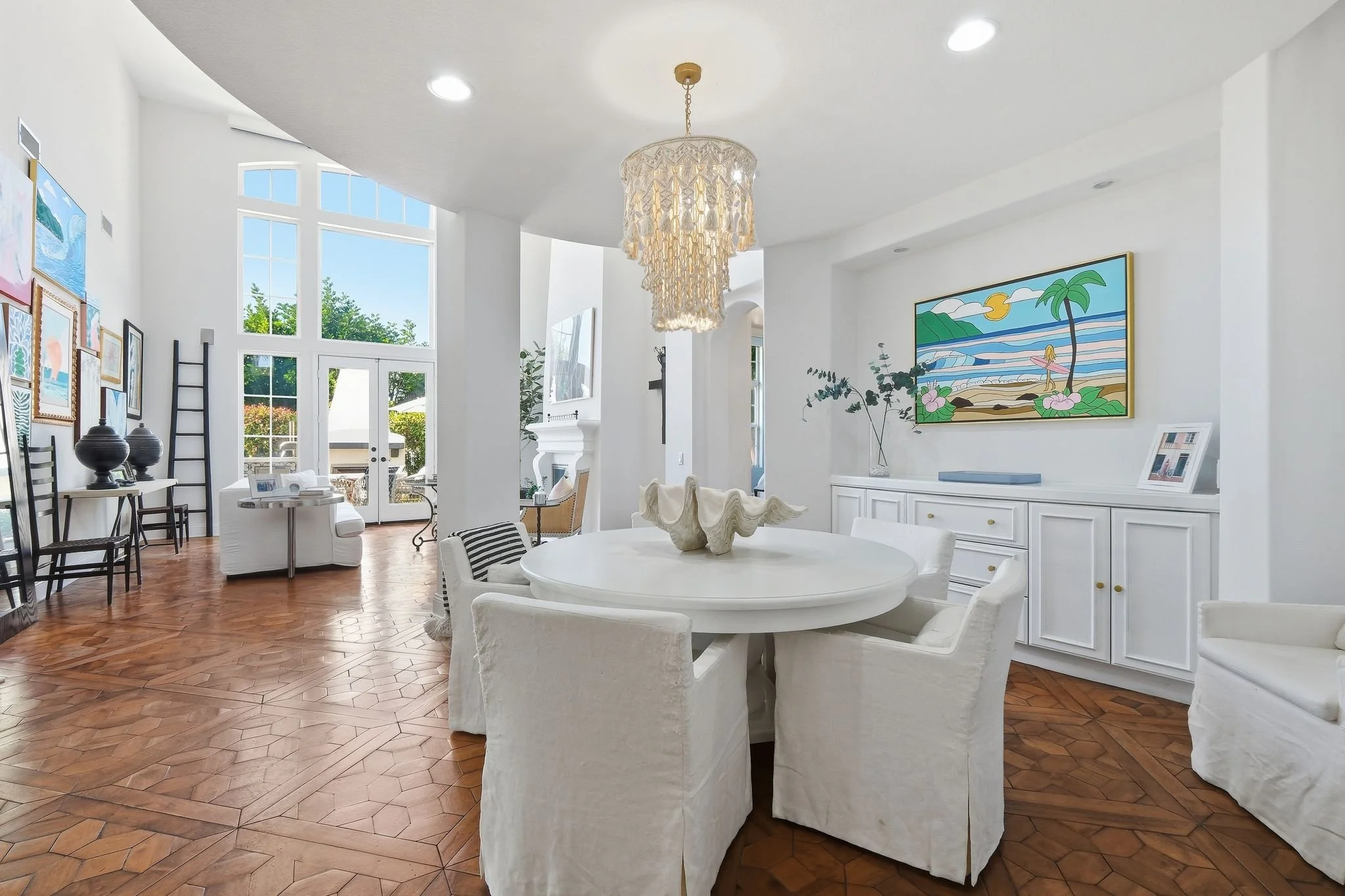 Bright open living and dining area in Carlsbad, San Diego with white walls, large windows, and warm wood floors. Features a round white dining table with six slipcovered chairs, a chandelier, and curated artwork - high end design



