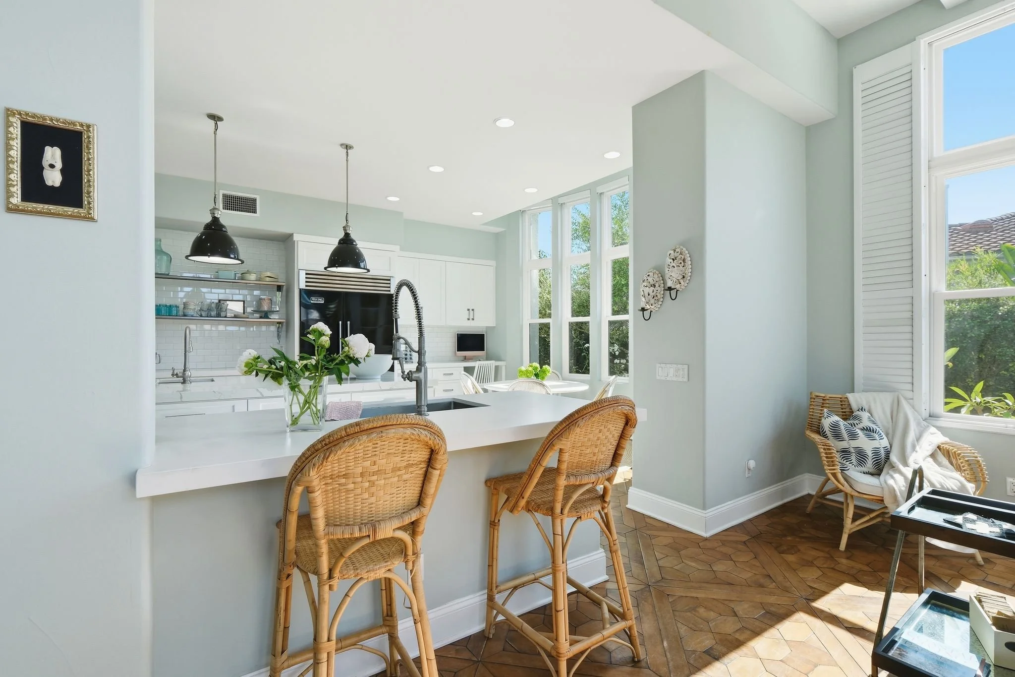 Bright modern kitchen with white cabinets, black appliances, and large windows, featuring a breakfast bar with wicker chairs and a small sitting area with a wicker chair and decorative pillows.