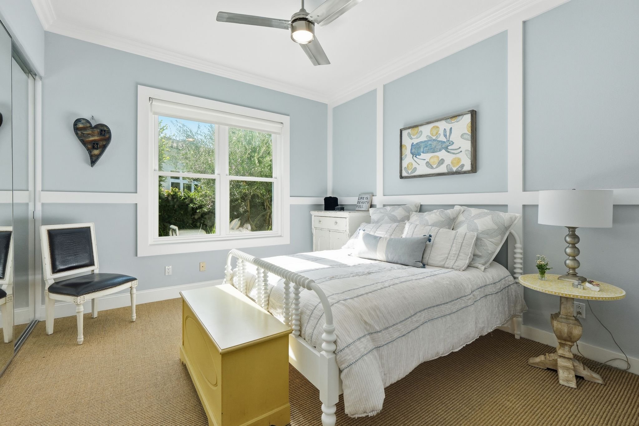 A bedroom with light blue walls, white trim, and a large window showing greenery outside. The bed has white bedding with gray and beige pillows. To the right of the bed is a rustic wooden nightstand with a white lamp and a small plant. On the wall ab