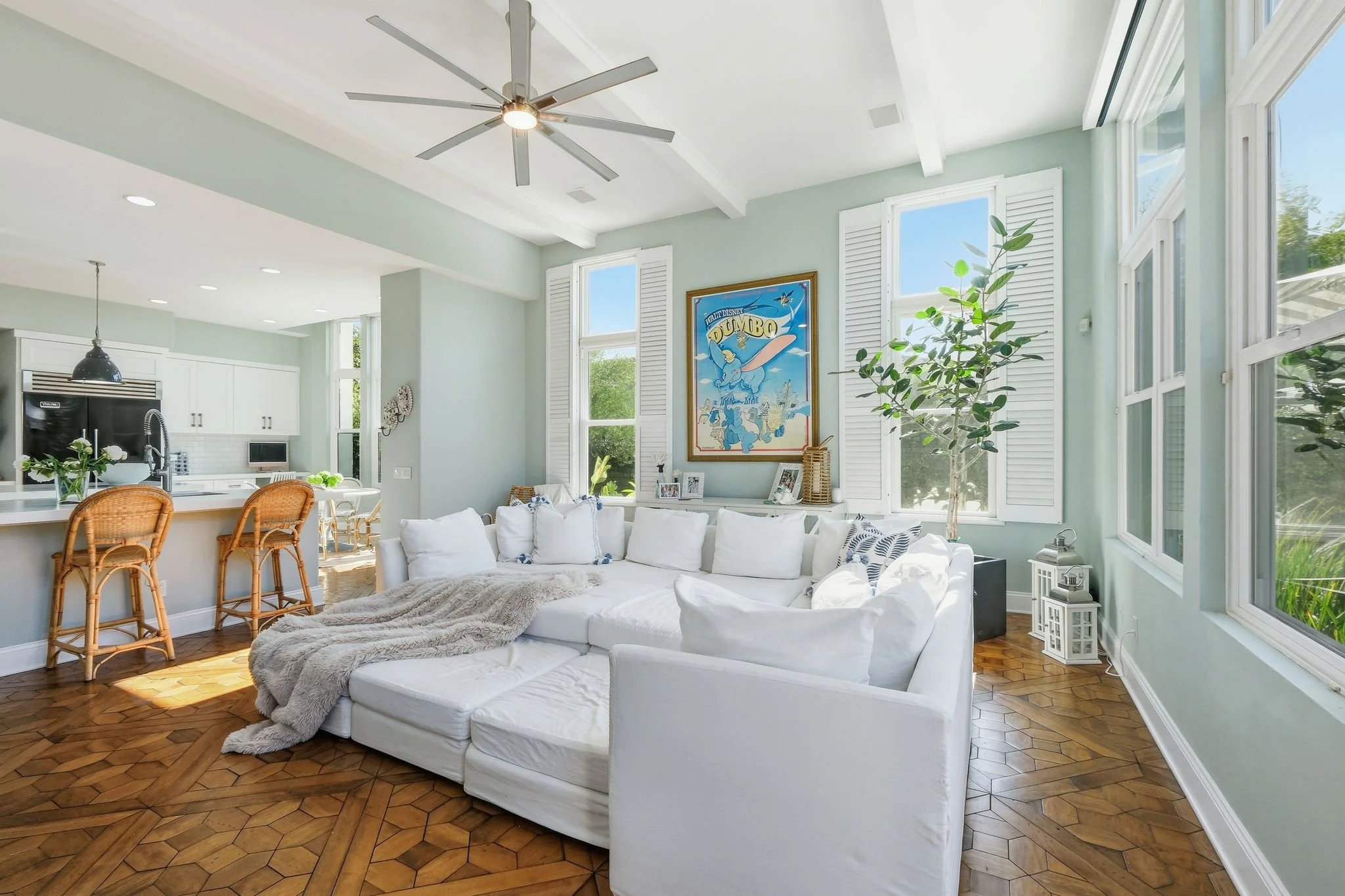 Living room with a white sectional sofa, wooden flooring, large windows, potted plant, and wall art of Dumbo.
