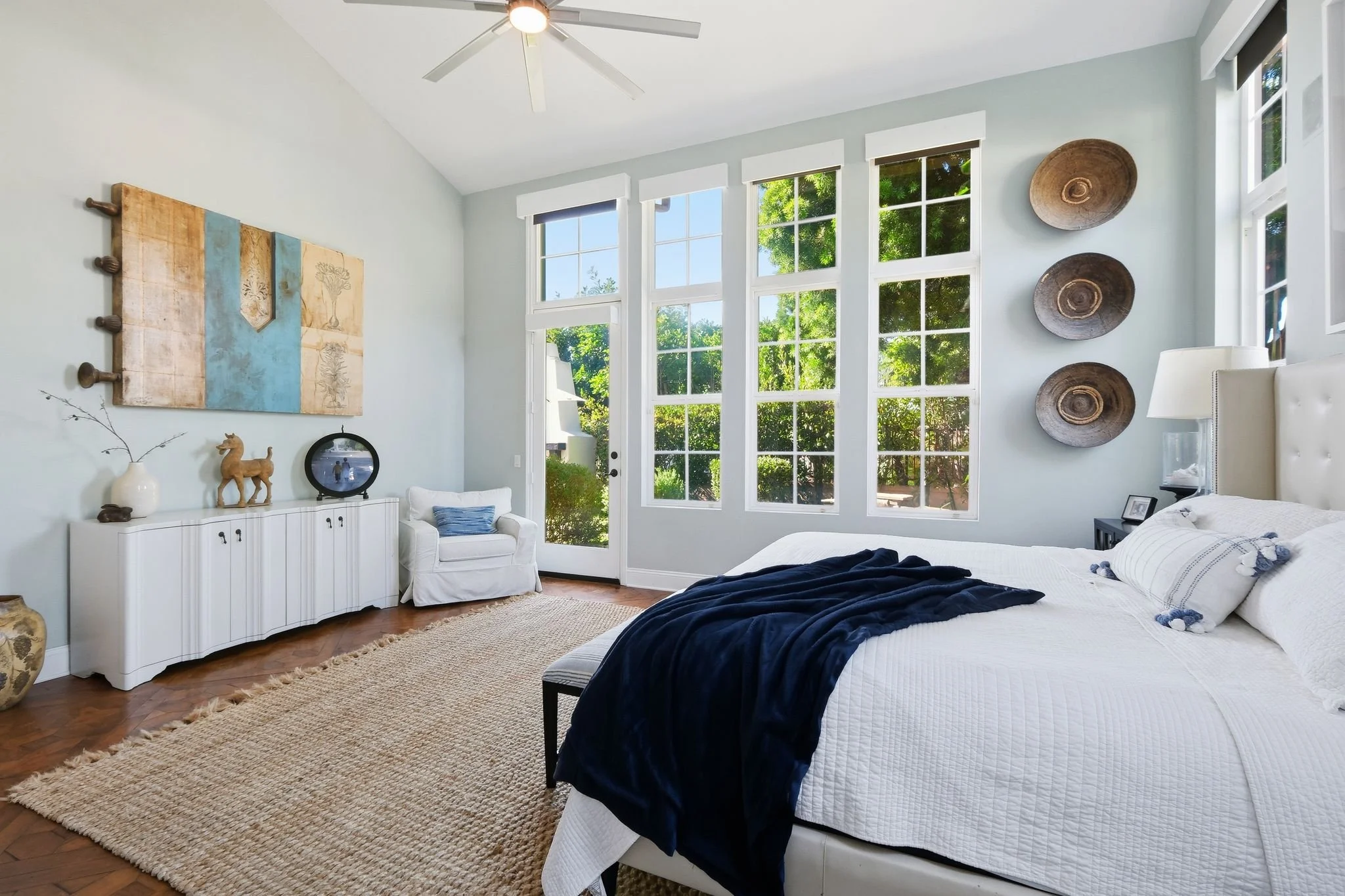 Bright bedroom with a large bed covered in white quilt and navy blanket, white upholstered headboard, nightstand with lamp, large windows with greenery outside, white armchair, textured rug, and decorative wall art.