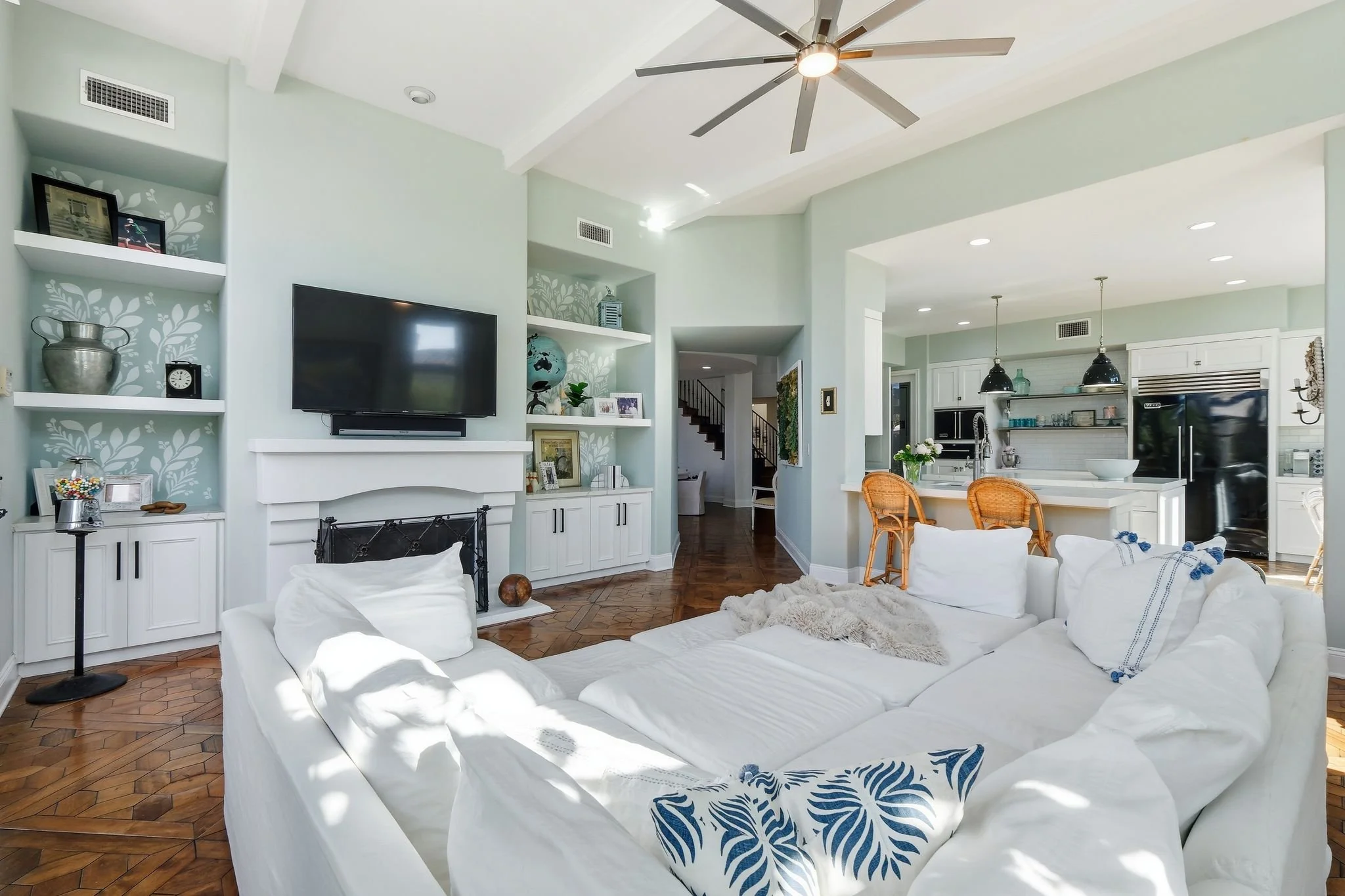 Bright living room with a white sectional sofa, wooden flooring, built-in shelves with decorative items, a TV above the fireplace, and an open kitchen with pendant lights and bar stools.
