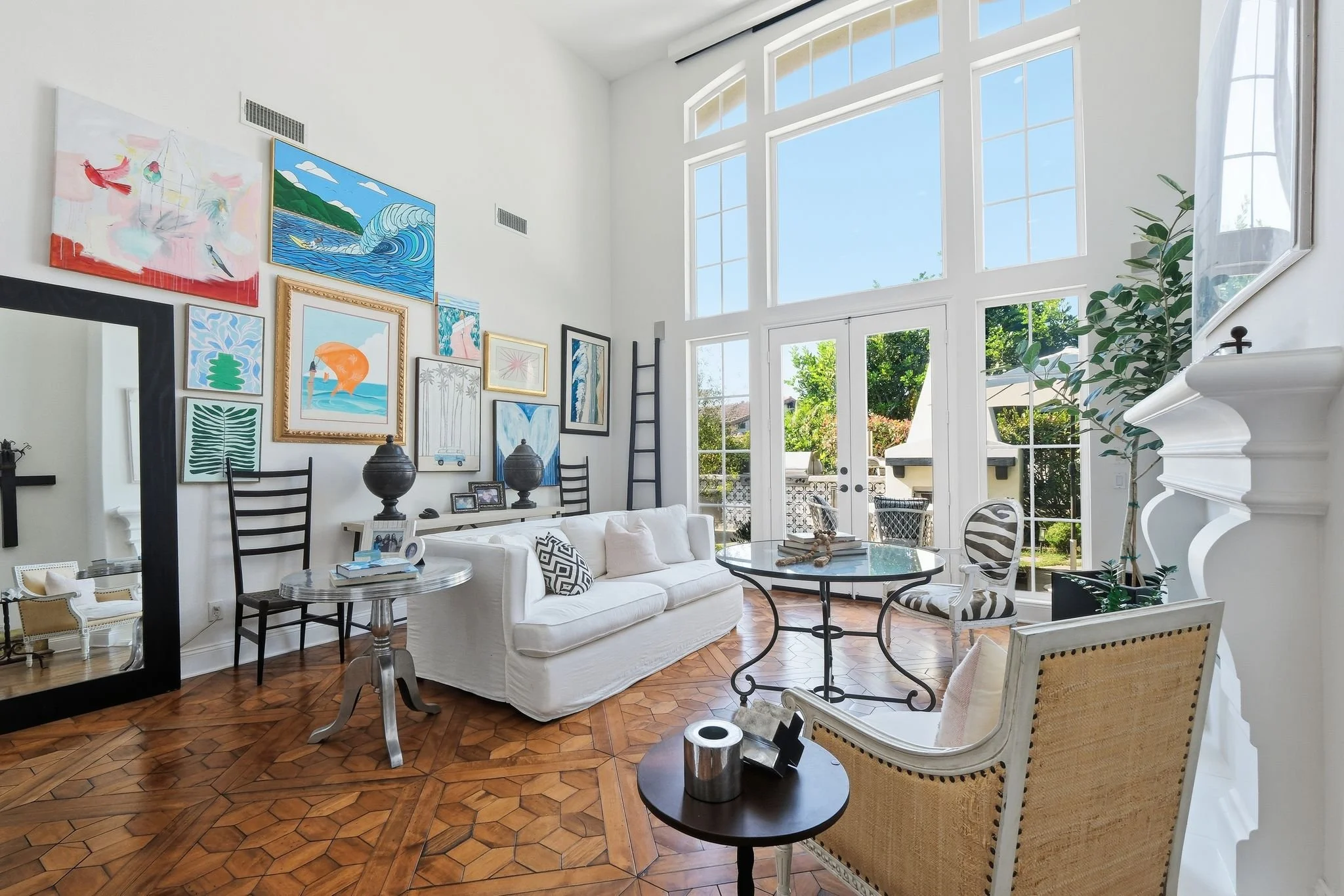 Bright, high-ceiling living room in Carlsbad, San Diego with large windows, hardwood floors, colorful wall art, a white sofa, layered seating, a round glass coffee table, and potted plants. A California-casual, eclectic space.